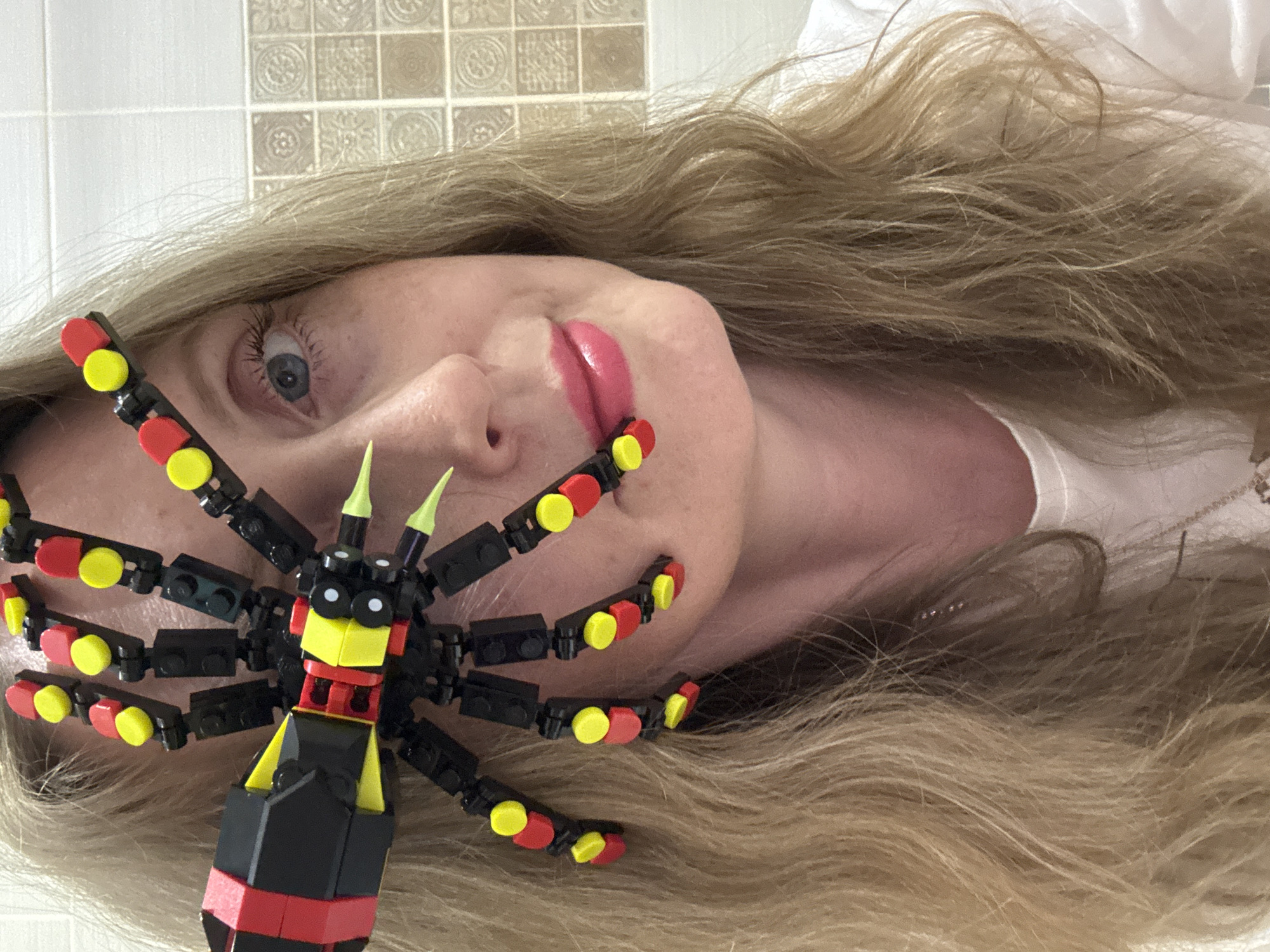 A woman with blonde hair is lying down, her head turned towards the camera. A Lego spider is positioned near her face. The background appears to be a tiled wall. The location in the city of Chaikovsky, Russia is not discernible from the image.