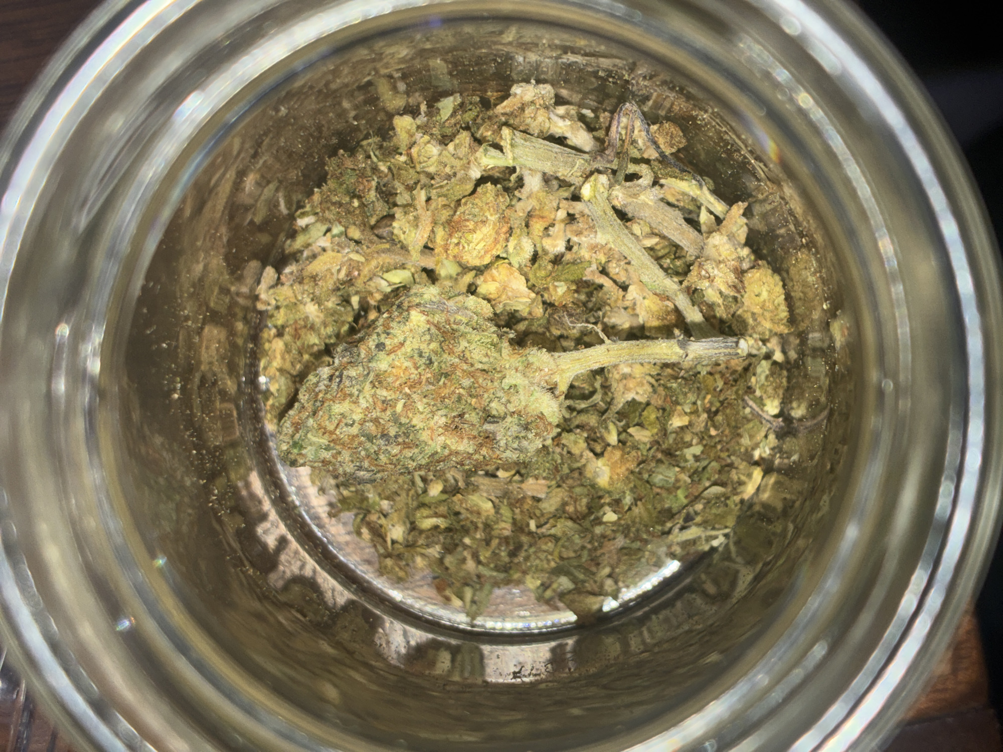 The image shows a close-up, overhead view of a glass jar filled with marijuana. The contents consist of a mix of green and brown plant material, including buds, stems, and leafy debris. The jar is clear, and the contents are visible through the glass. The surrounding environment is out of focus, but it appears to be a wooden surface. There is no information to suggest the image was taken in Huixquilucan de Degollado, Mexico.