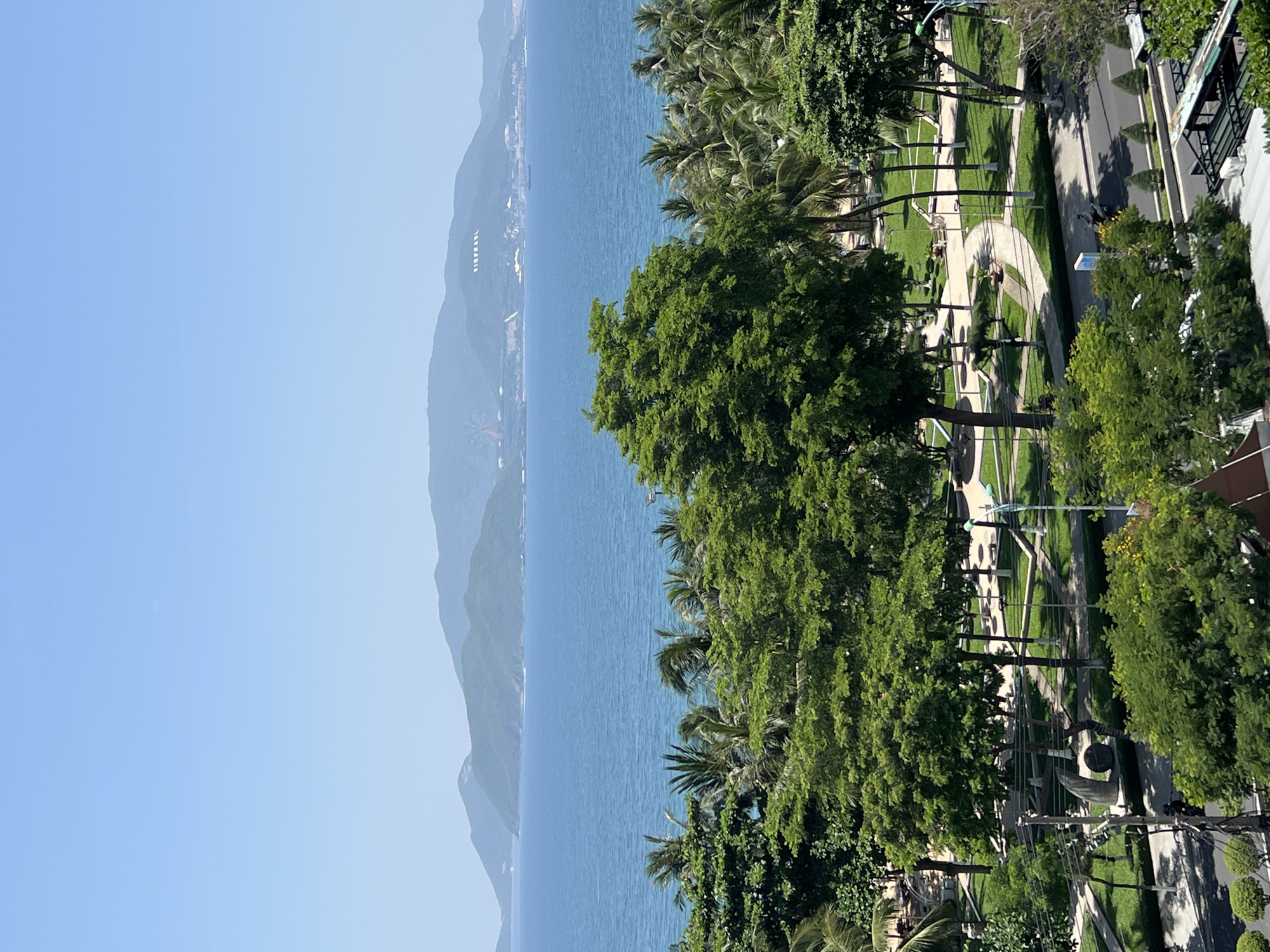 This is a high-angle, medium shot of a coastal city scene in Nha Trang, Vietnam, dominated by the bright blue ocean and lush green foliage under a clear sky. In the distance, a chain of hazy green mountains forms the horizon, with a faint inscription of white letters that appears to read "VINPEARL" on one of the hillsides. The sea is calm and stretches across the mid-ground of the image.
In the foreground and mid-ground, a vibrant park area is visible. This park features manicured green lawns, winding paved pathways, and numerous trees, including large deciduous trees with full canopies and tall palm trees. Several lampposts are dotted throughout the park. A road runs along the edge of the park, and on it, a few motorcycles and what appear to be street vendors are present. The overall impression is one of a bright, sunny day in a well-maintained urban coastal environment.