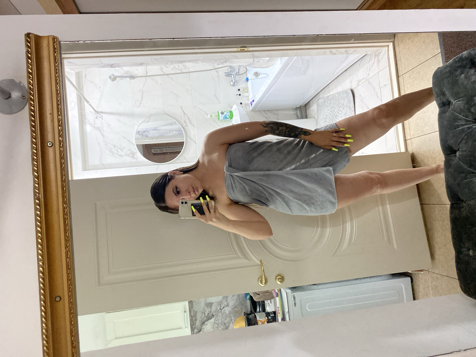 A woman stands in front of a mirror, taking a selfie. She is wrapped in a gray towel and her nails are painted a bright neon yellow. In the reflection, a bathroom with white marble tiles is visible, containing a bathtub and sink. To the left of the woman, a white washing machine and kitchen cabinets are seen. The overall environment suggests a residential setting, likely an apartment in Makhachkala, Russia.