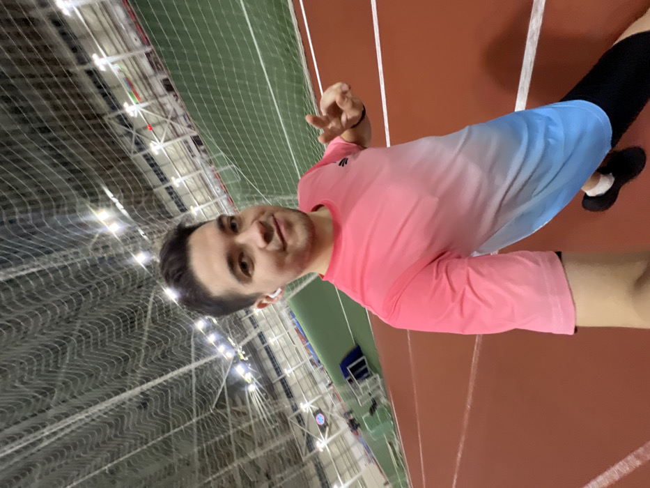 A person in a pink and blue shirt stands in a building with a green net ceiling. They are on a red running track, wearing black athletic bottoms and shoes. They are holding their hand up, making a hand gesture. The building has many lights.