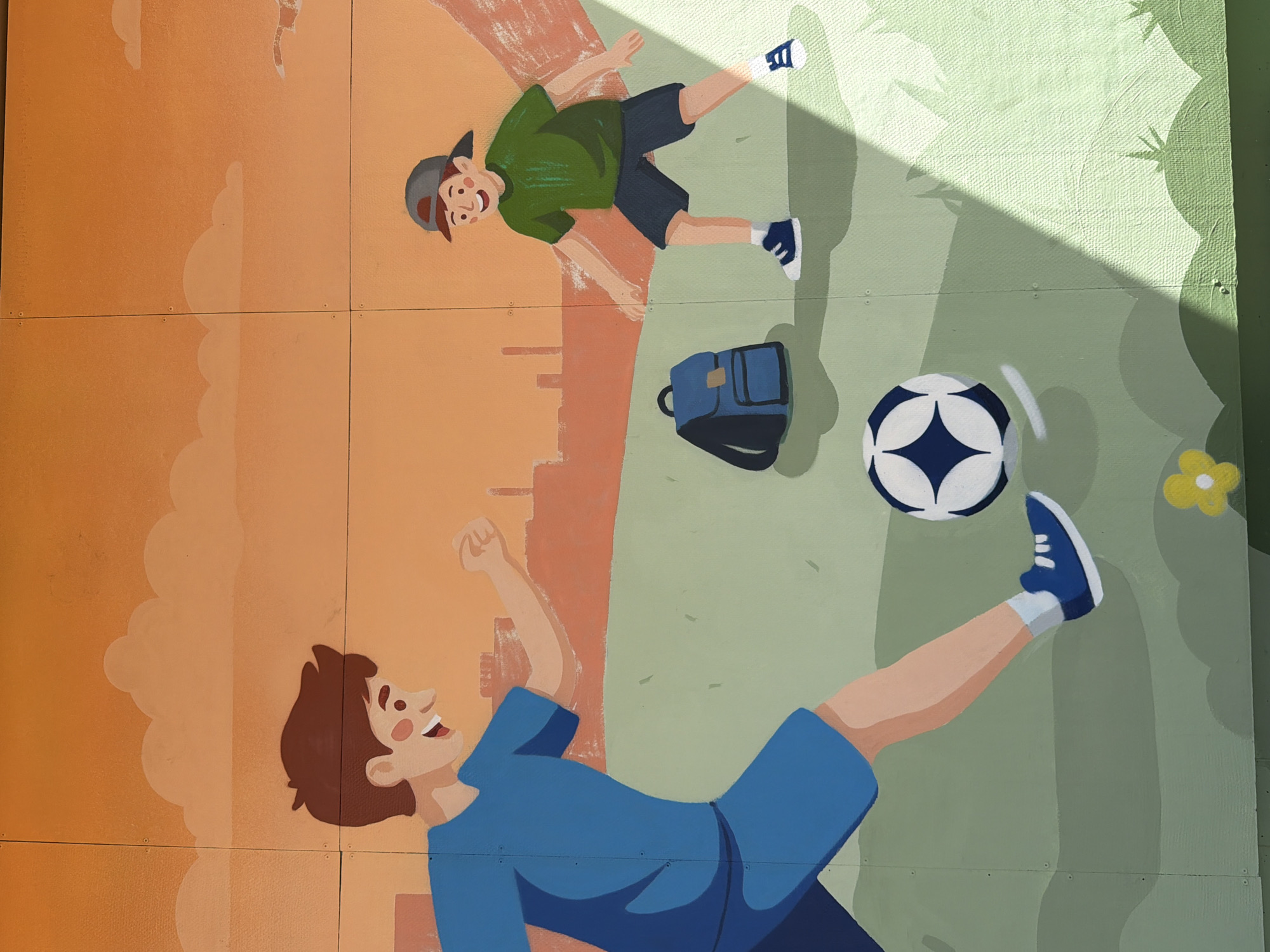 This is a mural painted on a building, depicting two young boys playing soccer. The boy on the left, wearing a blue shirt and shorts, is in motion, kicking the soccer ball. He has brown hair and is smiling broadly. The boy on the right, wearing a green t-shirt, dark shorts, and a baseball cap, is positioned further down the field and appears to be watching the ball. He also has brown hair and a smile. A blue backpack is visible on the grass between the two boys. The background of the mural shows a hazy orange sky with stylized clouds, and a silhouette of industrial buildings in the distance. The ground is depicted as rolling green hills with some yellow flowers. A shadow from an unseen object casts a diagonal line across the right side of the scene. The mural is painted on panels that are joined together. It is unclear from the image if this mural is located in Yekaterinburg, Russia.