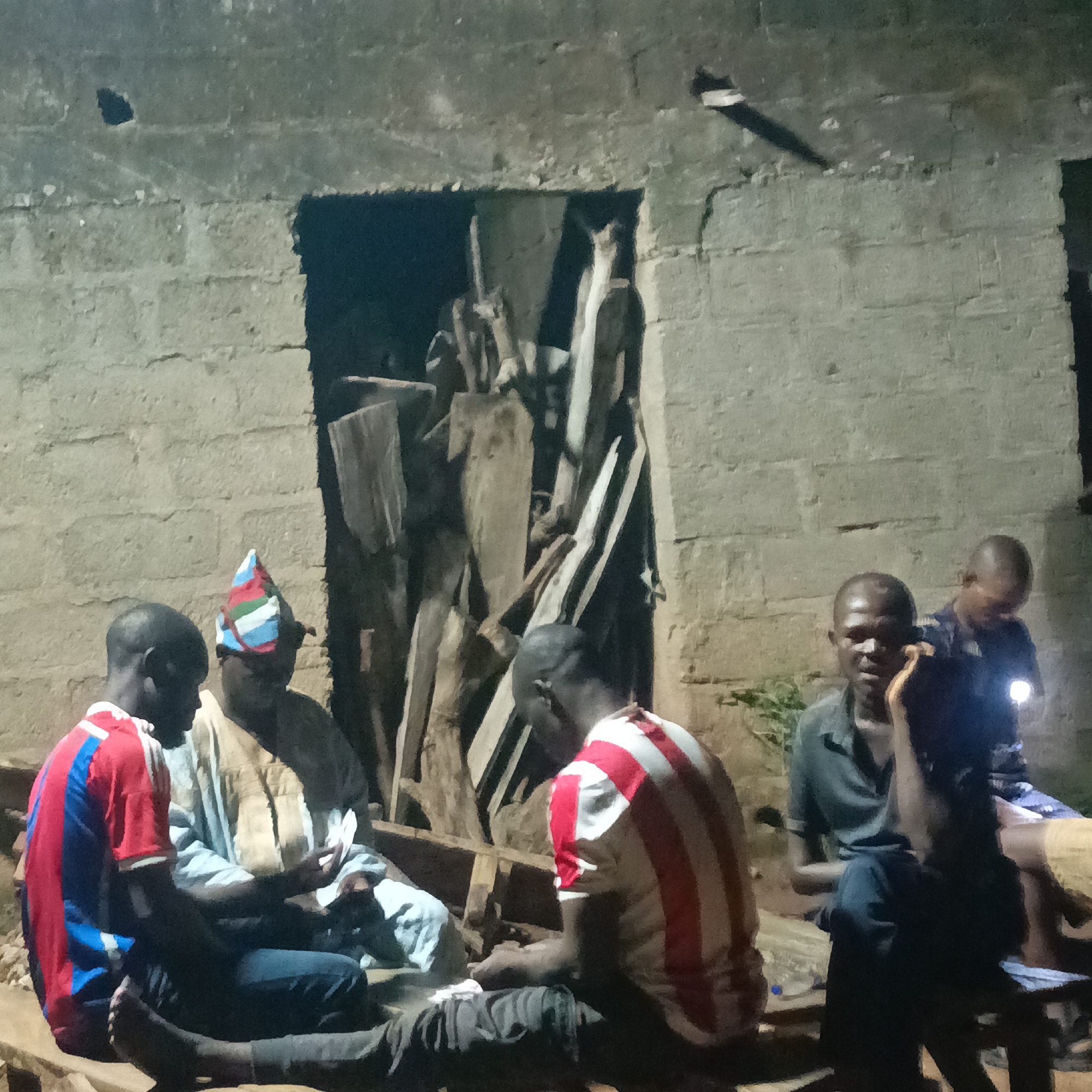 A group of five Black men are gathered outdoors at night, seated on what appear to be wooden benches and crates in front of a rough, unplastered concrete block wall with a dark, open doorway filled with stacked lumber. The lighting suggests a single light source off-camera, creating shadows. One man in the center, wearing a traditional brown robe and a colorful conical hat, is holding cards. To his left, a man in a red and blue striped shirt is looking at cards. To the right of the man in the robe, two other men are visible. One man in a green shirt is looking downwards, and another man further back, holding a light, is looking towards the camera. A fifth man is seated in the foreground to the right. The scene is likely in the city of Akwanga, Nigeria.