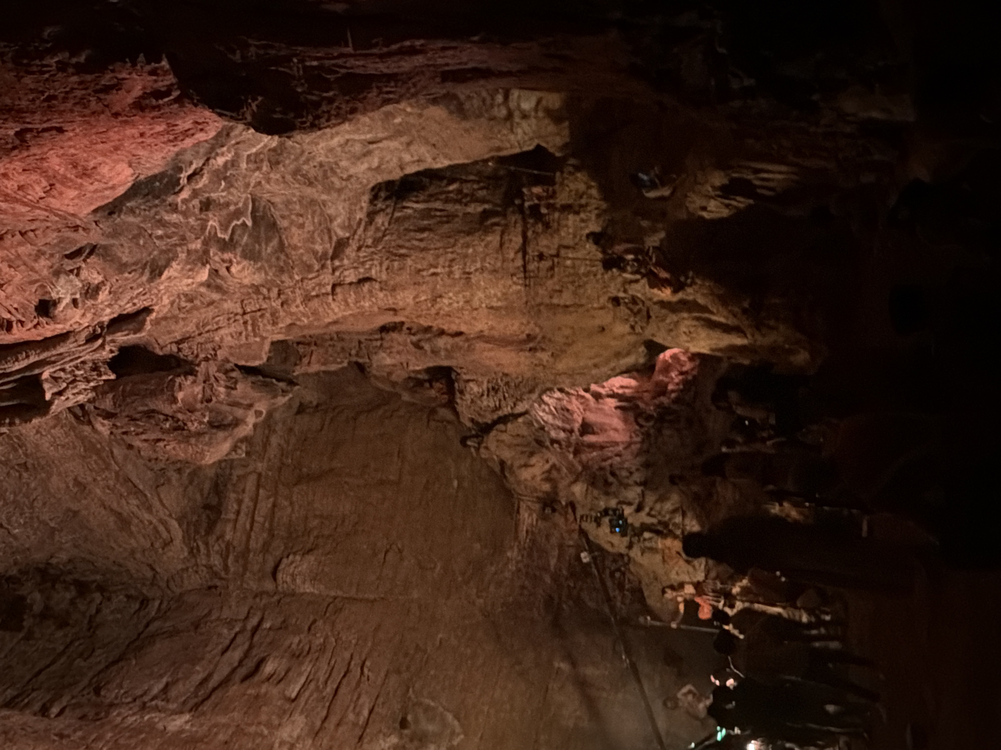 A group of people are gathered inside a large, dimly lit cave. The cave walls are rough and textured, with layers of rock formations illuminated by reddish-orange lighting. Some individuals are standing and looking around, while others appear to be walking or interacting. In the background, on a higher ledge, figures dressed in what appear to be costumes are posed, possibly as part of a performance or display. The overall impression is that of a tourist or event taking place within a natural cave environment. The specific location of Guttapadu, India, is not discernible from the visual information present in the image.