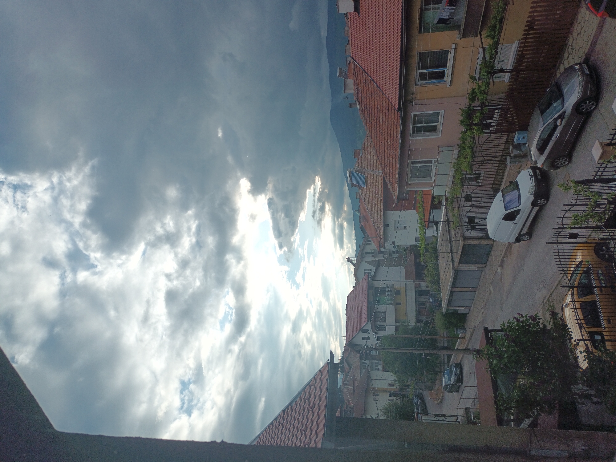 View of a neighborhood with buildings, cars, and cloudy sky. The buildings have red tile roofs and are various colors. A few cars are parked on the side of the street. The sky is filled with dark, cloudy skies with rays of sunlight breaking through.