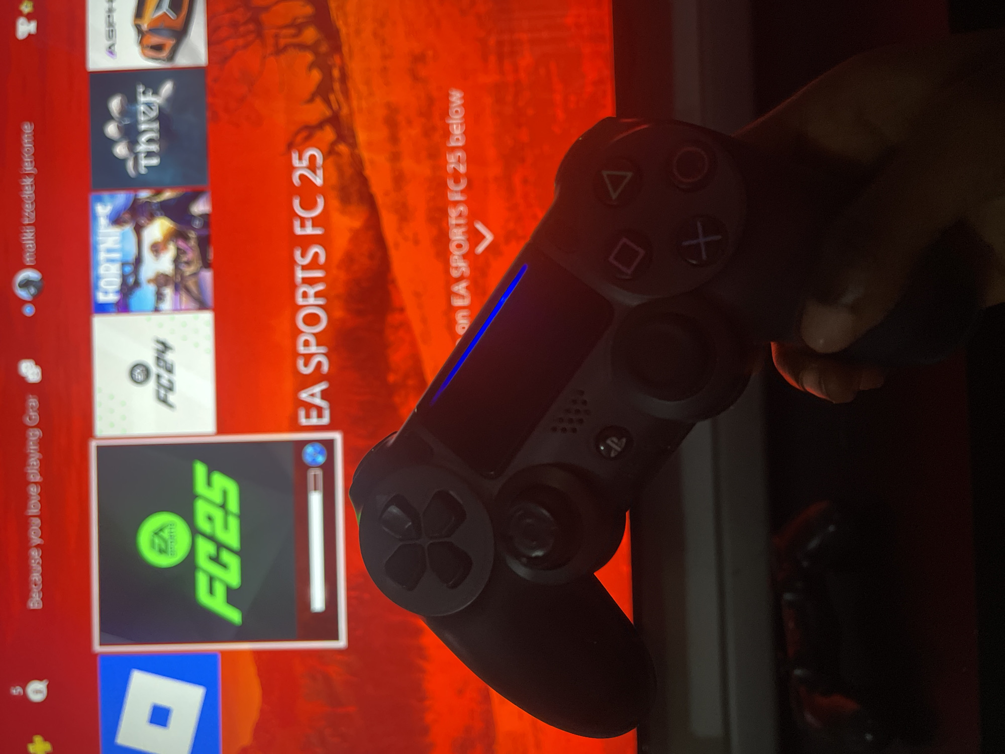 A person is holding a black PlayStation 4 DualShock controller in front of a television screen. The screen displays the PlayStation main menu, with various game tiles visible. The selected game tile is for "EA SPORTS FC 25". Other games visible include "FORTNITE," "FC 24", "ASPHALT", "Thief", and "Roblox". The PlayStation user is "malki tzedek jerome". The scene is presumed to be indoors, likely in a residential setting in Ado, Nigeria, but there is no further information to confirm the location.
