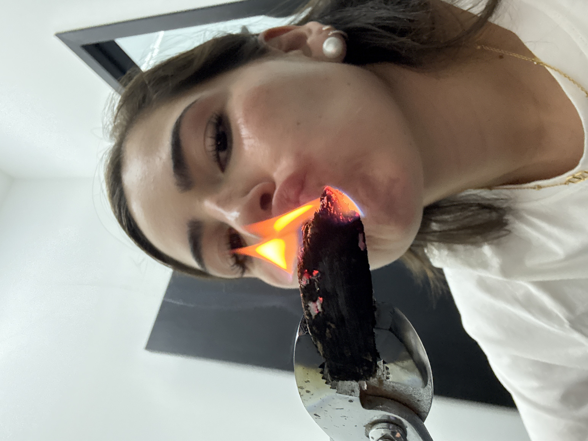 A woman with brown hair and fair skin is holding a lit piece of what appears to be charred wood or charcoal in a pair of metal tongs. The flaming end of the object is near her open mouth. She is wearing a white shirt and a pearl earring. The background includes a wall and a partially visible picture frame. The image was not taken in San Bernardino Tlaxcalancingo, Mexico.