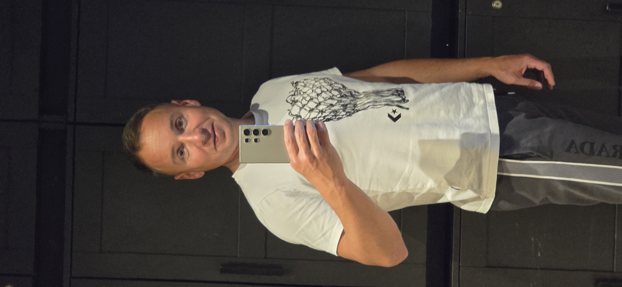 A man is taking a selfie indoors, presumably in a building in the city of Bar, Montenegro. He is holding a smartphone in his right hand. He is wearing a white t-shirt with a graphic design and grey sweatpants, which have a white stripe and a "PRADA" logo. The background features a dark cabinet-like structure.