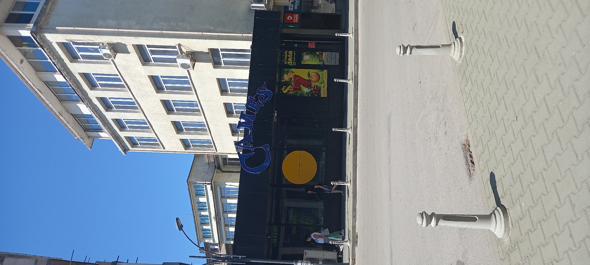 The image shows a street scene in Silistra, Bulgaria, dominated by the facade of a building with a dark ground floor and an upper structure with multiple windows. A large, illuminated blue sign on the dark ground floor reads "GAMES". Below the sign, there are various advertisements and posters, including one featuring the number "7" and fruit, and another with text in Bulgarian.
Two women are visible walking on the sidewalk in front of the building. One woman on the left, wearing a white top and a striped skirt, carries a bag. The other woman, on the right, is wearing a dark top and denim shorts, and appears to be wearing headphones. Several concrete bollards line the sidewalk along the street. The street itself is paved, and a section of cobblestone pavement is visible in the foreground. The sky above is clear and blue, indicating a sunny day. The overall environment suggests an urban setting with commercial establishments.