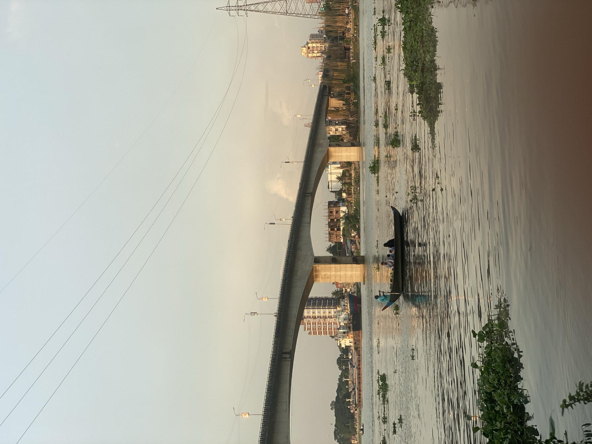 A serene river scene unfolds in Tarabo, Bangladesh, under a pale, overcast sky. A long, elevated concrete bridge with multiple spans dominates the upper half of the frame, supported by robust concrete pillars. Numerous streetlights are affixed to the bridge's underside, casting faint illumination. On the far bank, a cityscape is visible, featuring several multi-story buildings of varying architectural styles, including one with a distinct red facade. A prominent electricity pylon stands tall on the right side of the image.
The river itself is a muted brown color, reflecting the sky and the surrounding structures. The water's surface is rippled, disturbed by a gentle current. Scattered across the river are patches of lush green water hyacinths, some floating freely and others forming dense mats.
In the foreground, a narrow, dark wooden boat navigates the water, carrying four individuals. One person, dressed in a teal shirt, stands at the stern, seemingly steering the boat with a long pole. Three other passengers are seated in the boat.
Further in the distance, a large cargo ship with a blue hull is anchored, alongside other smaller vessels and boats near the shore. The overall atmosphere is calm and tranquil, suggesting a late afternoon or early evening setting. The environment appears to be an urban waterfront, characterized by both natural elements like the river and aquatic plants, and man-made structures such as the bridge and buildings.