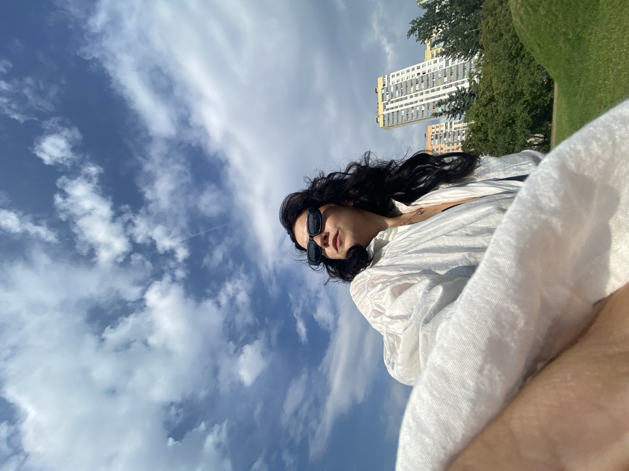 A low-angle, outdoor, eye-level shot features a woman with dark, wavy hair wearing black sunglasses and a loose white shirt. She is looking to her left, and her chest and a portion of her leg are visible in the foreground, covered by the white shirt. Behind her, a cluster of trees separates her from a cityscape, which includes tall apartment buildings with balconies. The sky above is blue with scattered white clouds. The scene is set on a grassy knoll.
