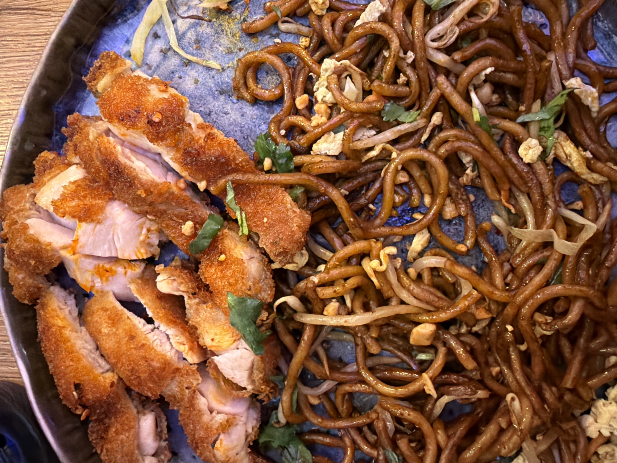 The image displays a close-up, overhead view of a prepared meal served on an irregularly shaped, dark blue ceramic plate with a dark, raised rim. The dish is composed of two primary elements: breaded fried chicken and stir-fried noodles.
In the upper section of the plate, multiple pieces of golden-brown, crispy breaded chicken are arranged. The chicken has been segmented, revealing the cooked white meat interior. Green herb fragments, consistent with cilantro, are scattered on and around the chicken pieces, which also show traces of reddish-orange seasoning on their breading.
The lower two-thirds of the plate is occupied by a quantity of dark brown, thin, curly stir-fried noodles. These noodles are intermingled with translucent, thinly sliced vegetables (likely onions or bean sprouts), small yellowish fragments resembling scrambled egg, and small, irregular, light-brown pieces, possibly chopped nuts or fried garlic. Additional green herb fragments are distributed throughout the noodle mixture.
The plate rests on a visible wooden surface, and a dark, unidentified object is partially visible in the upper left corner of the frame. The scene is illuminated by ambient indoor lighting. The specific location within Paris, France, is not ascertainable from the visual information provided.