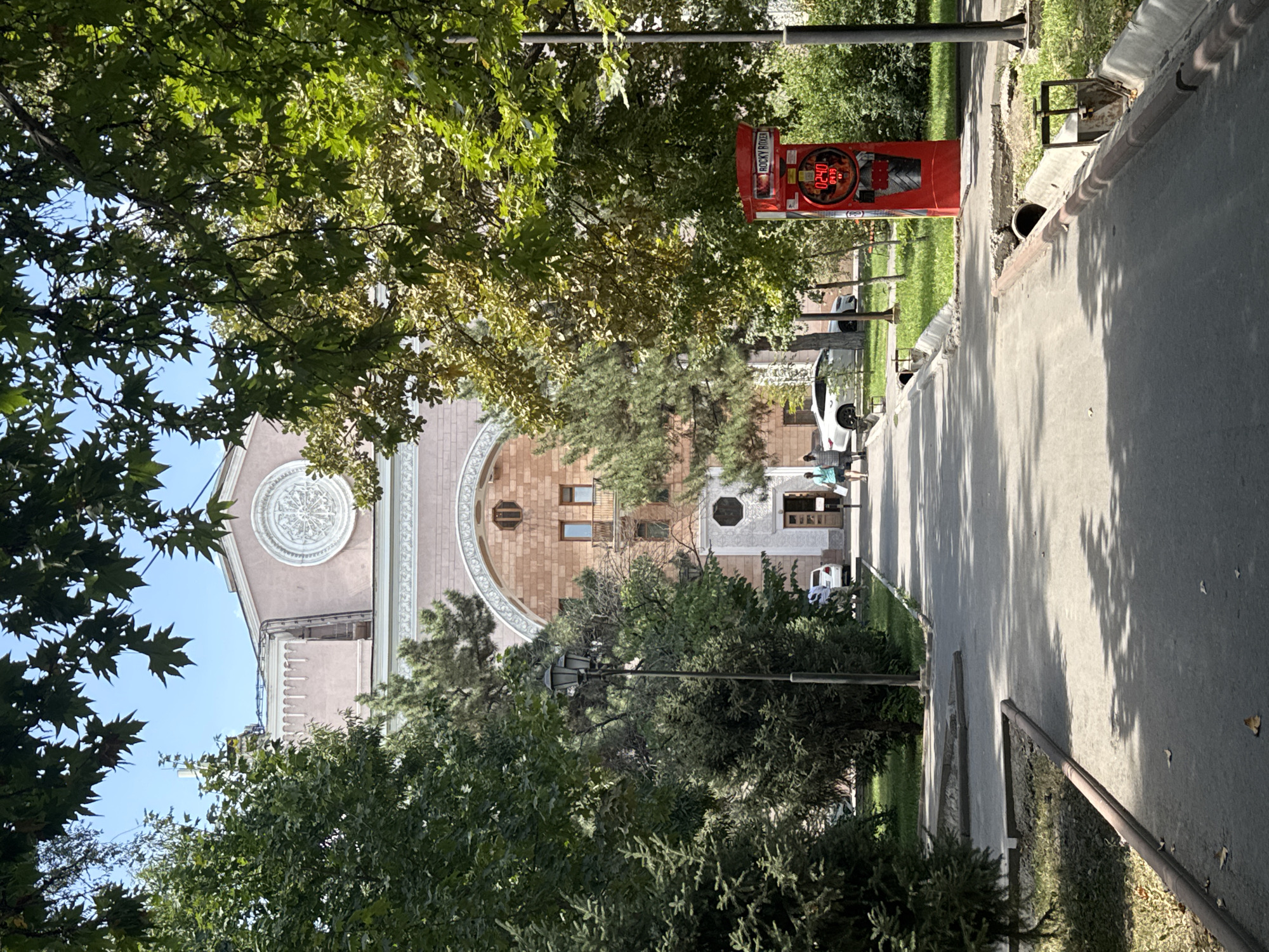 The image depicts an outdoor scene with a building in the background and trees lining a pathway in the foreground. The building is light pink with arched entrances and ornate architectural details, including a circular medallion on its facade. A paved pathway leads towards the building, with two individuals walking away from the camera on the right side of the path. To the right of the pathway, there is a bright red, upright object with a digital display showing "02.40" and "0435," accompanied by text that reads "ROCKY BOXER." A street lamp stands on the left side of the pathway, and various trees with green foliage are present throughout the scene, casting shadows on the ground. The environment appears to be a park or a public space within a city. The overall impression is a sunny day in a well-maintained urban area.