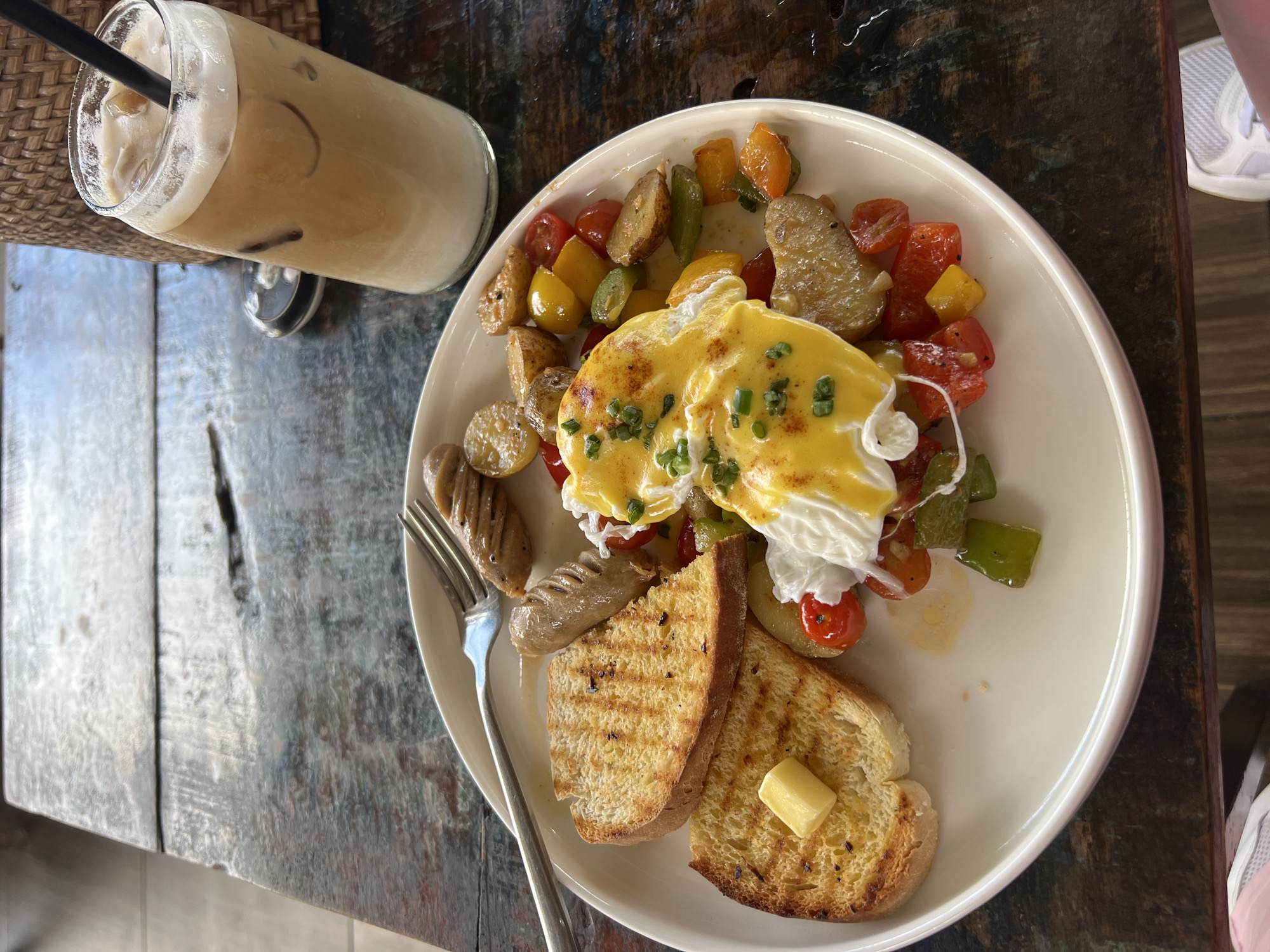 The image displays a bright, overhead view of a breakfast or brunch meal and an iced coffee, set on a rustic wooden table, likely in North Kuta, Indonesia.
The central subject is a white, round plate holding a substantial meal. Two poached eggs, generously covered in a creamy yellow hollandaise sauce and sprinkled with chopped chives, are the focal point. Surrounding the eggs are roasted potato pieces, vibrant sautéed vegetables including red and yellow cherry tomatoes and green bell peppers, and two grilled sausages. Two slices of grilled bread, one topped with a small pat of butter, are also on the plate, with a silver fork resting beside the sausages.
To the left of the plate, a tall glass of iced coffee, possibly an iced latte, stands with a black straw. The drink shows distinct layers of light brown coffee and white foam on top. Part of a woven rattan mat is visible beneath the glass.
The table surface is made of dark, distressed wooden planks, showcasing a textured and worn appearance. Natural light illuminates the scene, suggesting it is daytime. The edge of a white athletic shoe is visible in the top and bottom right corners, indicating a person is seated at the table. There is no visible text in the image.