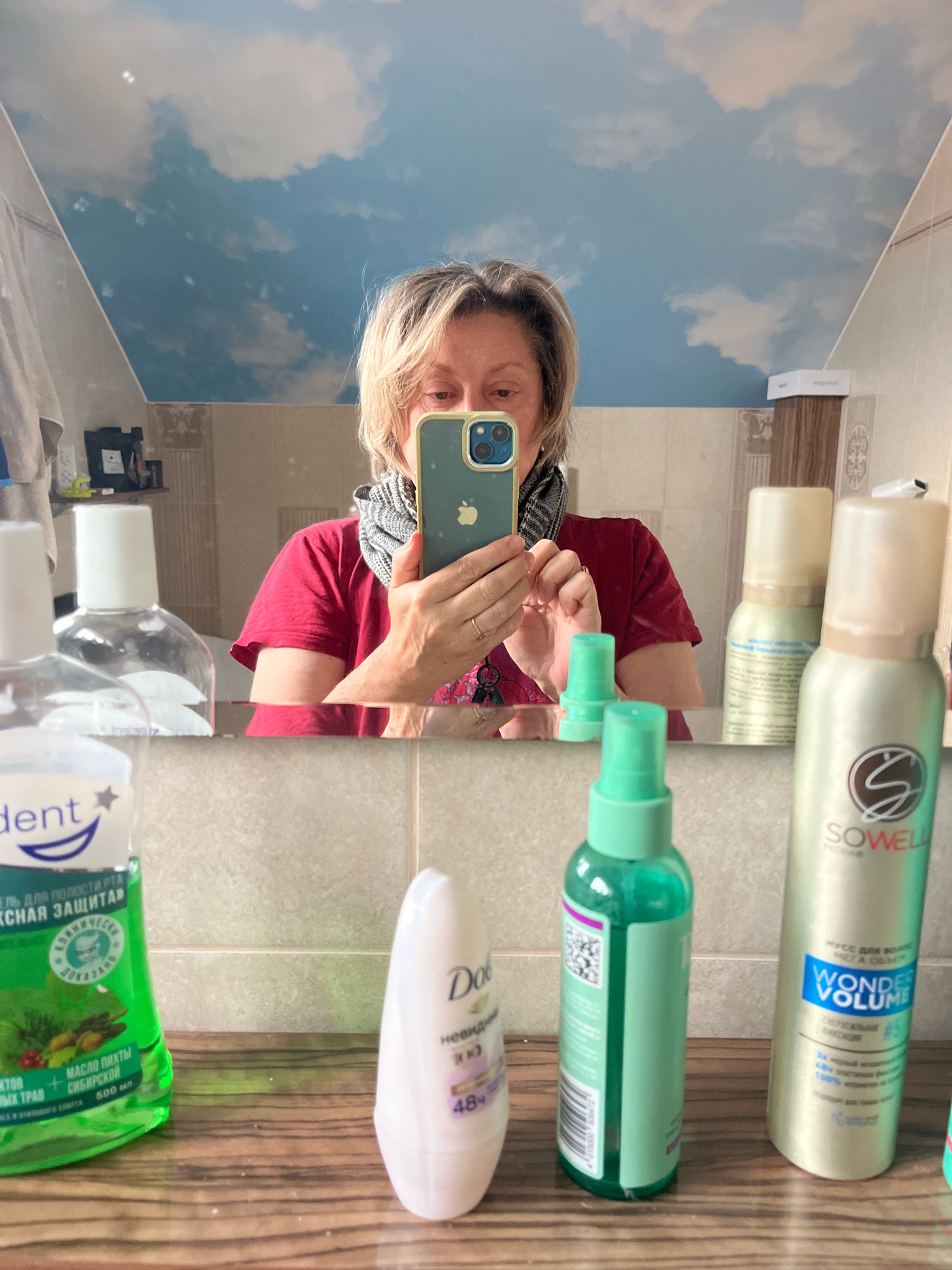 A woman is taking a mirror selfie in a bathroom. She is holding a smartphone in her right hand and is wearing a red shirt and a patterned scarf. The mirror reflects her image and the bathroom environment. In the foreground, on a wooden countertop, are several personal care products. To the left is a green bottle of mouthwash, followed by a white roll-on deodorant, a green spray bottle, and two aerosol cans. The bathroom walls are tiled, and the ceiling features a cloud-patterned wallpaper. The scene is consistent with a residential bathroom. There is no specific information within the image to indicate a location in Vladimirovka, Russia.