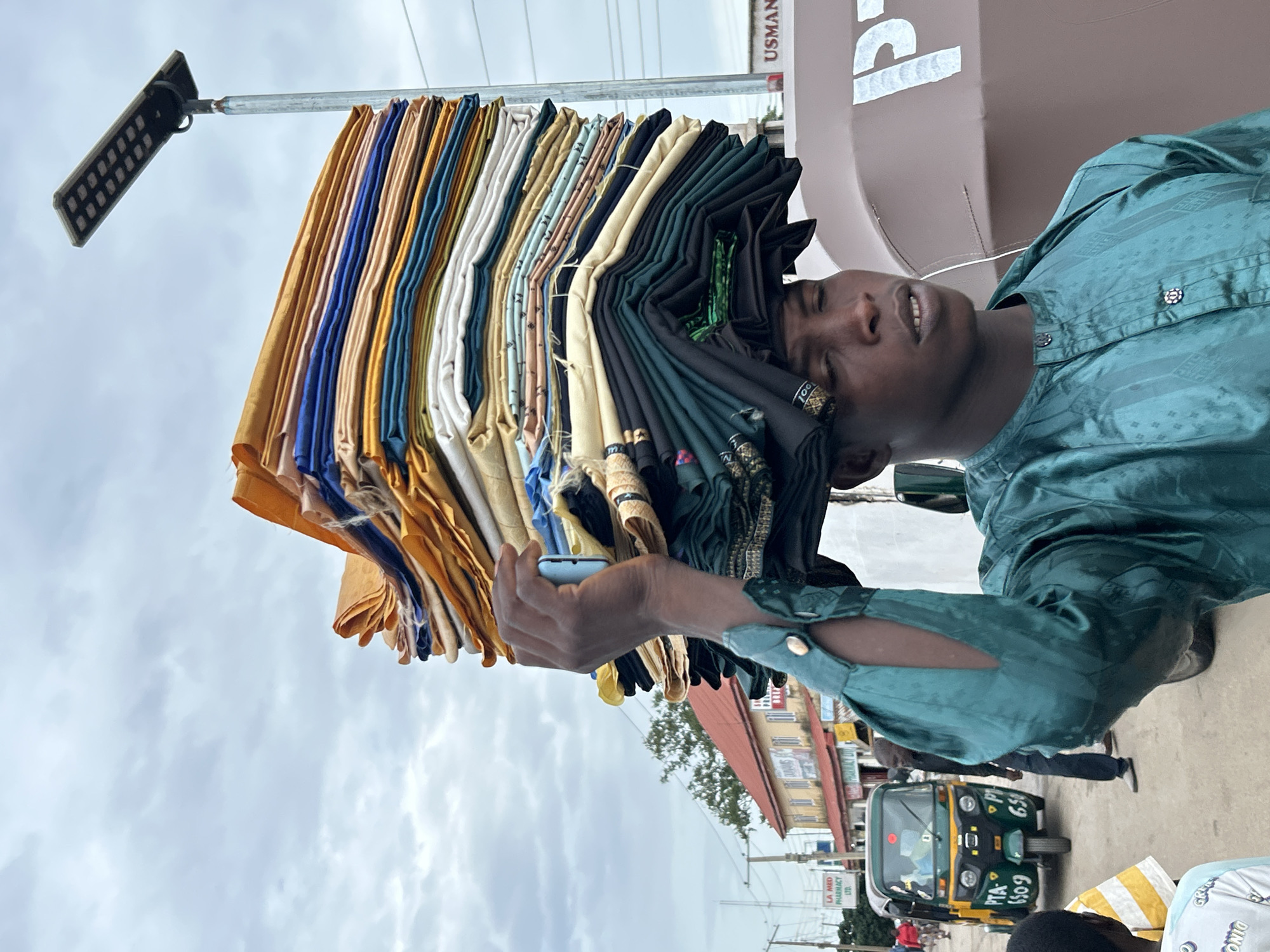 A man is depicted in a medium shot, facing slightly right, carrying a large, multicolored stack of folded fabrics on his head. He wears a teal-colored long-sleeved tunic with button closures and intricate patterns. His right arm is raised, with his hand supporting the base of the fabric stack, and he holds a light blue smartphone between his thumb and index finger. A black and gold patterned fabric or head wrap is visible beneath the main stack on his head. His eyes are slightly closed or downcast, and his mouth is slightly open.
The stack of fabrics consists of numerous layers in various solid colors, including gold, yellow, blue, teal, white, beige, green, and black, with some patterned pieces integrated. The stack is significantly taller than the man's head.
The background reveals an urban street scene under an overcast sky. To the left, a green and yellow motorized tricycle taxi, commonly known as a "keke Napep," is visible with the numbers "PTA 6509" and "650" printed on its side. Beyond it, a building with a red roof and yellow walls has signs, including "LA MED PHARMACY LTD" and "DUNAMIS." Several indistinct figures are also present on the street.
To the right of the man, a tall pole supports a modern solar-powered LED streetlight. Below the streetlight, a large, cylindrical brown water tank is visible, with the letters "PT" partially discernible on its side, and the name "USMAN" above it on a white sign. The ground appears to be asphalt or concrete. This scene is located in a commercial or residential area within the city of Jos, Nigeria.