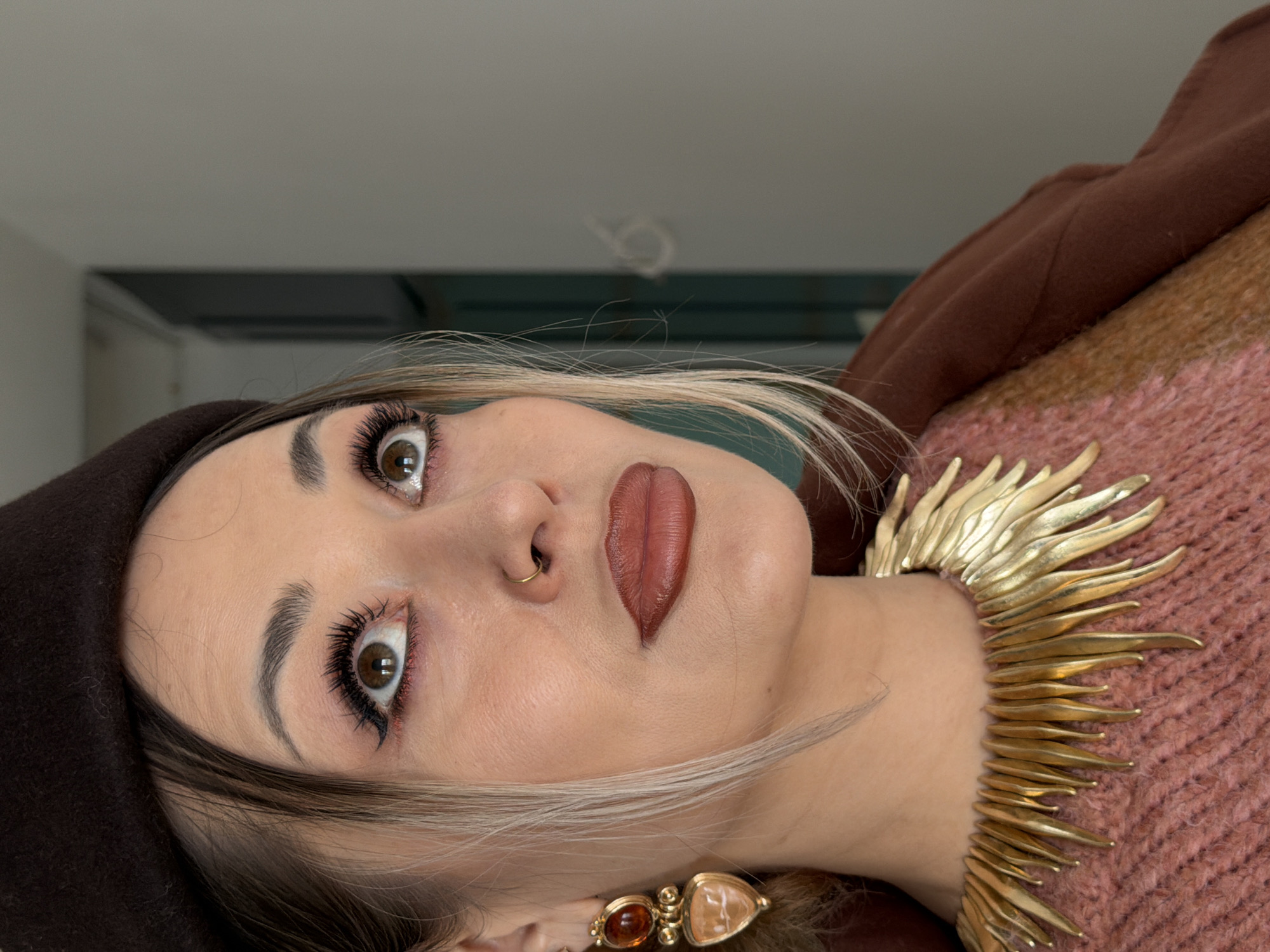 A woman is shown in a close-up shot, appearing to be indoors. She is wearing a brown beret and a pink sweater, accessorized with a gold necklace and earrings. The woman has fair skin, brown eyes with prominent eyelashes, and brown lipstick. A nose ring is also visible. The background is a neutral color with a hint of teal. There are no indications that this image was taken in Krasnoyarsk, Russia.