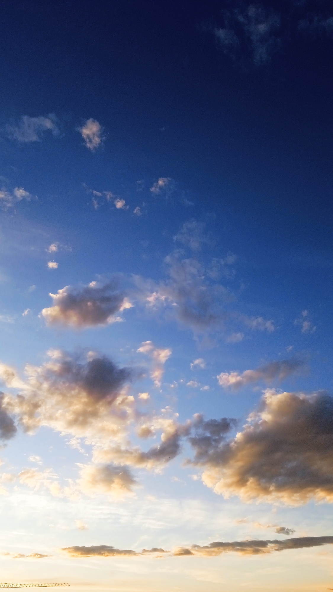 A vertical, medium-shot photograph captures a vast expanse of the sky, transitioning from deep, rich blue at the top to lighter hues and wispy clouds towards the bottom. The upper portion of the image is a gradient of dark blue, indicating twilight or a deep daytime sky. Scattered throughout the mid-section are small, white clouds, some appearing fluffy and others more stretched. As the eye moves down, the clouds become more prominent and are tinged with warm, golden-orange light, suggesting the presence of the sun just above or below the horizon. These larger, more dramatic clouds are a mix of light and shadow, with some appearing dark and heavy, while others are illuminated from behind. The lower part of the image shows a very bright, almost white band of light, likely the horizon, with subtle, horizontal cloud formations. A thin, dark, horizontal structure resembling a bridge or railing is visible at the very bottom edge of the frame, partially obscured by the bright light. The overall atmosphere is serene and expansive, with a focus on the natural beauty of the sky at dusk or dawn. There are no people or animals visible in the image, and the environment is solely the sky and a hint of a man-made structure.