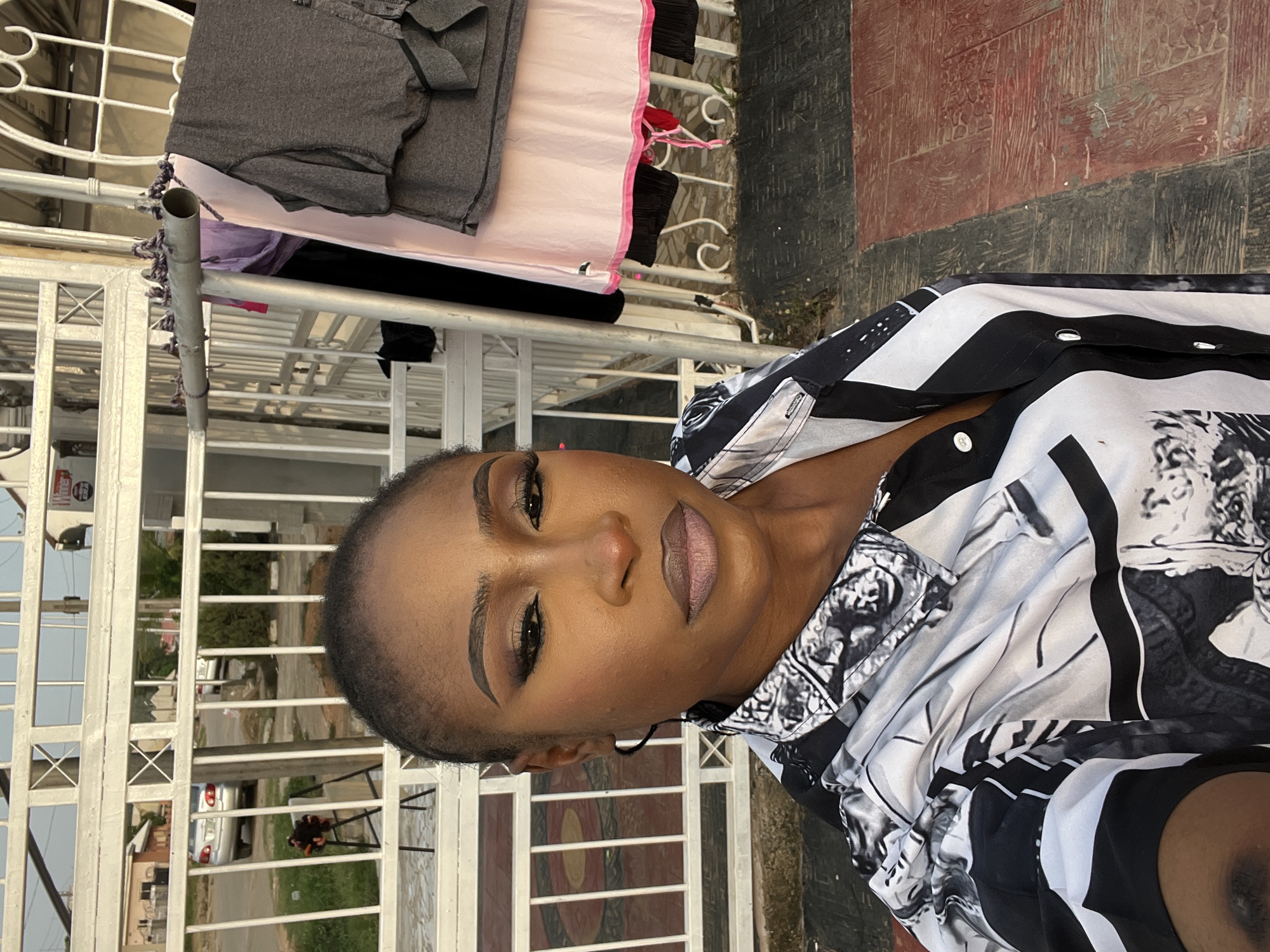 An African woman is taking a selfie outdoors in Abuja, Nigeria. She is wearing a black and white patterned shirt and has on makeup, including eyeshadow and lipstick. She is standing in front of a white metal fence. Clothes are hanging on a clothesline in the background. A street with cars and houses is visible behind the fence. The ground is paved with red and black tiles.