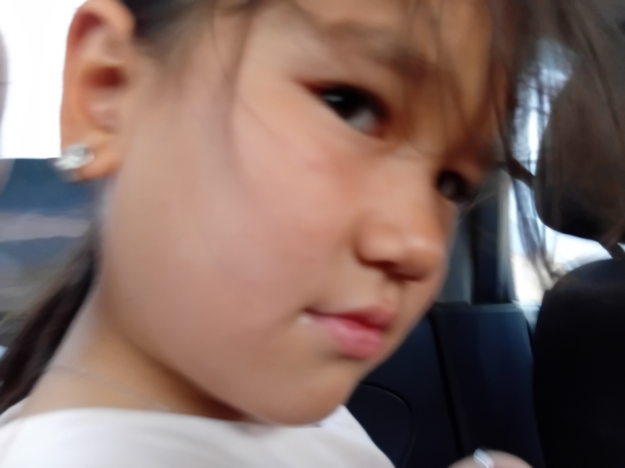 The media file shows a close-up, blurry, eye-level shot of a young girl's face. She has dark hair that is partially obscuring her face and a prominent ear with a small, sparkling stud earring. Her skin tone is fair, and her lips are slightly parted, revealing a hint of teeth. The background is dark and out of focus, suggesting an indoor setting, possibly a car, with hints of light and what might be seats. The overall lighting appears to be natural, possibly daylight, judging by the brightness on her skin and the diffused light in the background. The girl's expression is difficult to discern due to the blurriness, but she seems to be looking directly at the camera. There is no visible text in the image. The image's shallow depth of field keeps the focus primarily on the girl's face, particularly her eye and ear.