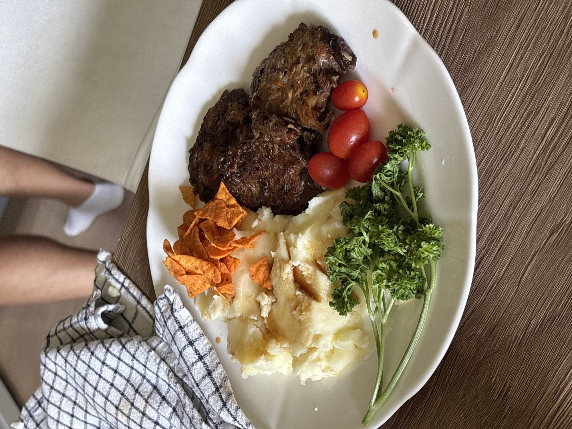 An overhead medium shot shows a white plate with food on a wooden table. A checkered cloth is draped on the left side of the plate, with a person's legs visible in the background.
The plate contains:
- Mashed potatoes drizzled with gravy.
- A pile of orange-colored chips.
- Several cooked patties, possibly burgers or meatloaf.
- A sprig of parsley.
- Four cherry tomatoes.
The food is arranged around the center of the plate. The mashed potatoes and chips are on the left, with the meat patties and tomatoes on the right. The parsley is at the bottom left.
The environment appears to be indoors, with soft lighting. The wooden table has a visible grain pattern. The person in the background is wearing white socks.
The image does not provide any information or context that would allow for determining the location in the city of Choeng Thale, Thailand.