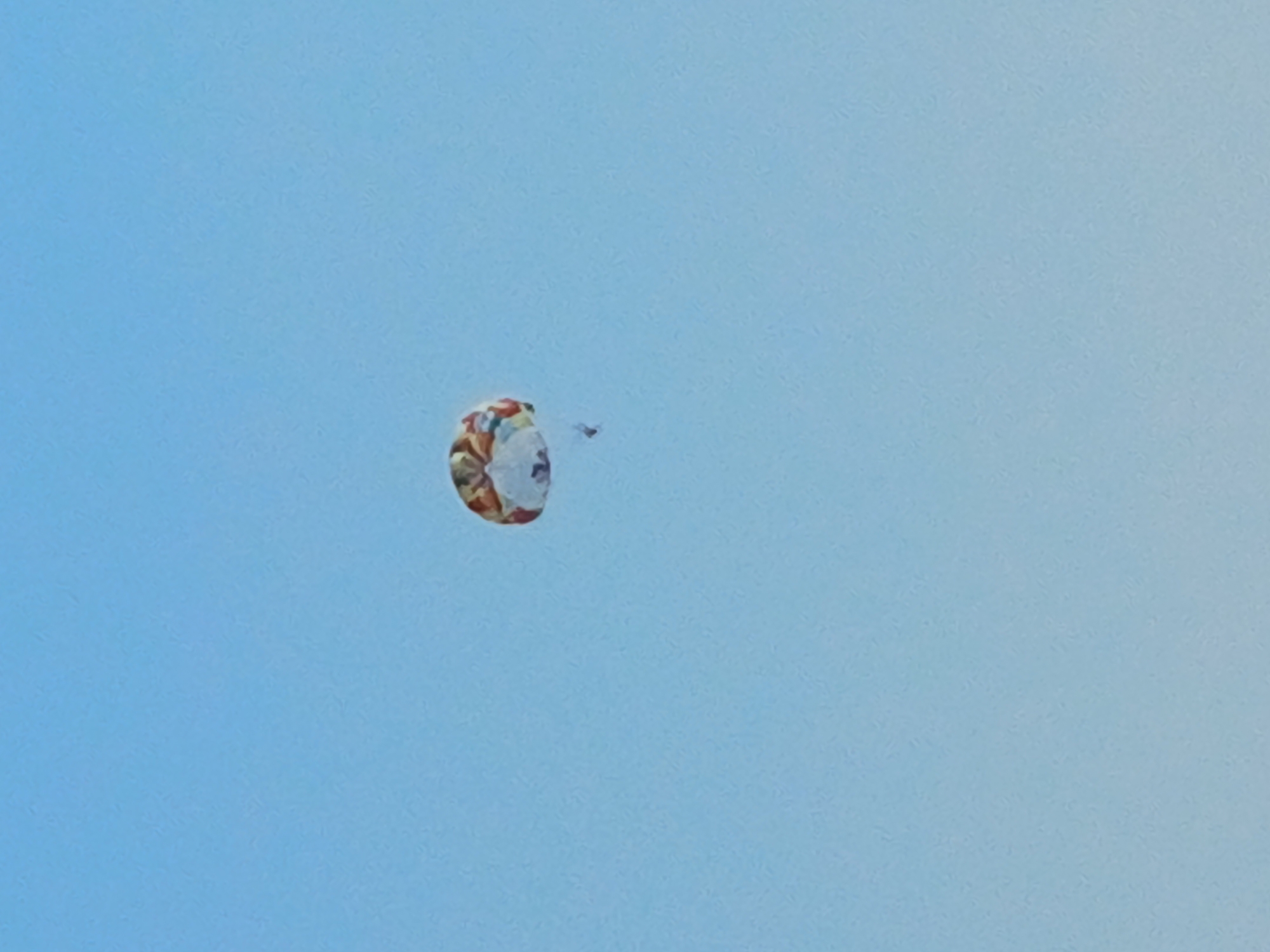 A colorful, circular parachute descends against a clear blue sky. A person is attached to the parachute by lines, but they are very small and difficult to discern. The parachute itself has an intricate pattern of orange, yellow, white, and blue colors. The image appears to be taken on a bright, clear day. The location is identified as Sochi, Russia.