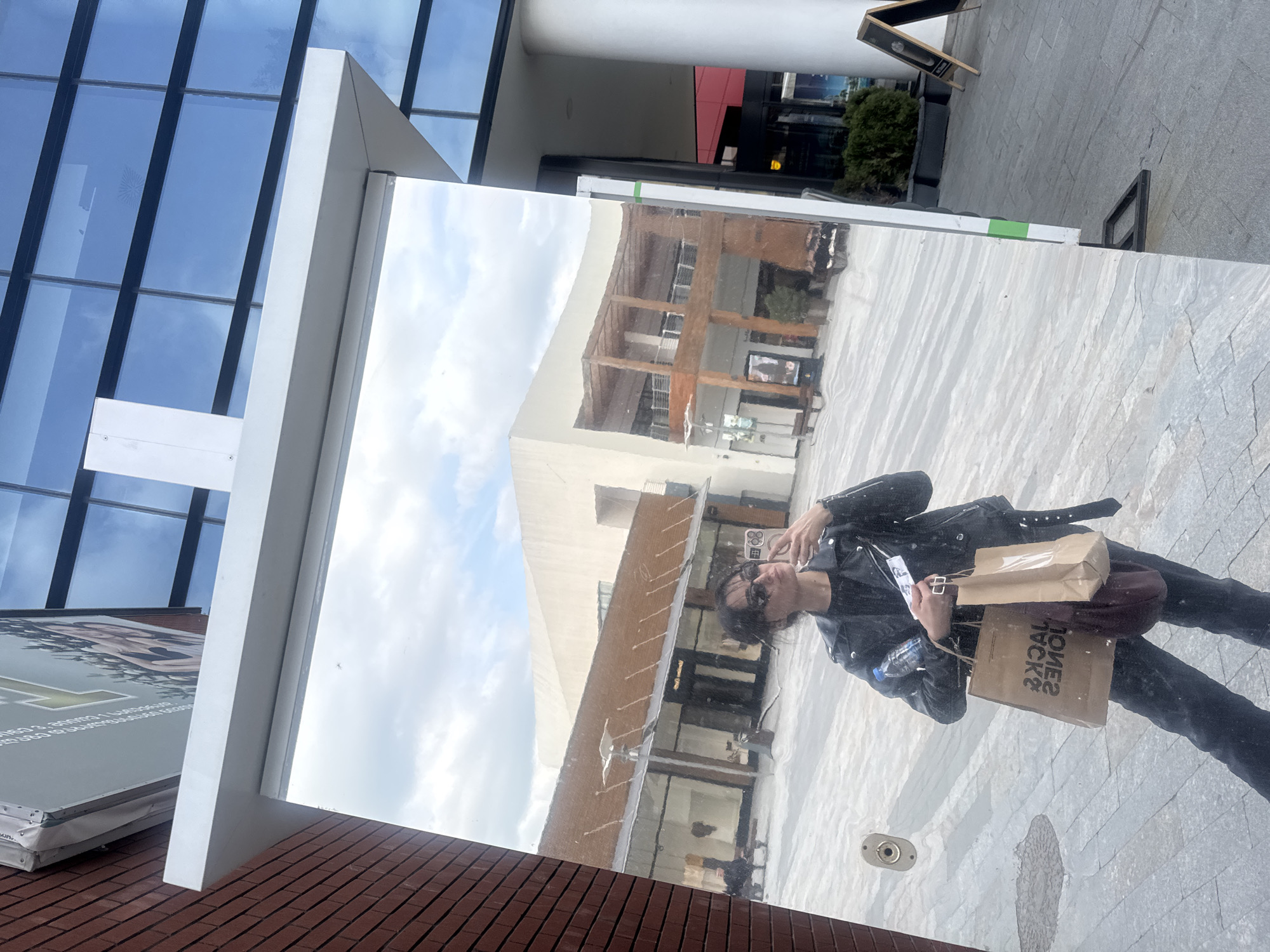 An individual takes a mirror selfie in an outdoor urban setting, likely a modern shopping area in Tbilisi, Georgia. The person, who appears to be a young adult, is dressed in a black leather jacket, dark pants, and sunglasses, and is holding two brown paper shopping bags, one prominently featuring the reversed text "JACK & JONES." A dark shoulder bag and a water bottle are also visible.
The selfie is captured in a large, vertical reflective surface or mirror, which is bordered by white framing and set on a red brick base. The reflection shows modern buildings with clean lines, glass, and wooden accents, as well as a paved ground and a cloudy sky. Beyond the mirror, the actual background features a modern building with blue-tinted glass windows, a white architectural element, and a grey paved walkway with a small green shrub and a partial view of a black A-frame sign. The overall scene is brightly lit, indicating daytime, under a partly cloudy sky. A circular metal fixture is visible on the paved ground near the bottom right of the mirror's reflection.