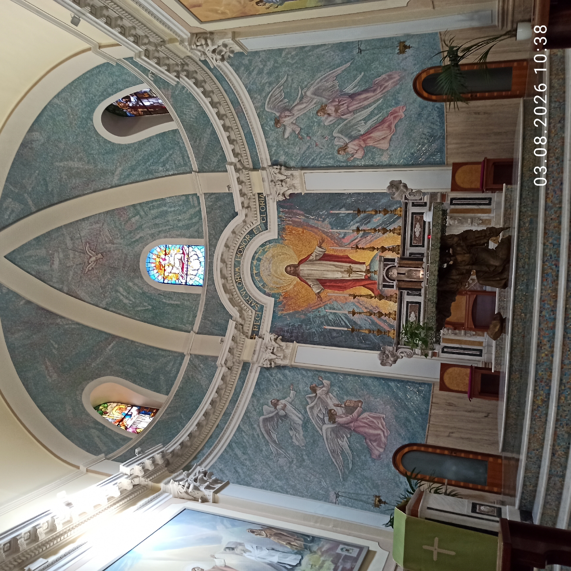 The image shows the interior of a church in Ruvo di Puglia, Italy, focusing on the altar area and the apse. The apse is decorated with a large mosaic of Jesus Christ with outstretched arms, set against a vibrant golden and orange radiant background. Above this mosaic, a Latin inscription reads "Regi saeculorum honor et gloria" (To the King of ages, honor and glory). The dome of the apse features a fresco of a dove, symbolizing the Holy Spirit, and a stained-glass window depicting a biblical scene.
Flanking the central mosaic are murals of angels with spread wings. On the left side of the altar, a mural depicts figures in white and ochre robes, possibly saints or biblical characters, illuminated by a bright light. On the right side, the mural continues with more depictions of angels.
The altar itself is a substantial structure with intricate carvings, featuring a sculpture of a kneeling winged figure supporting the table. Candles in ornate candelabra are placed on and around the altar. The steps leading up to the altar are adorned with colorful mosaic tiles.
To the left of the altar, a green lectern with a white cross is visible. Two wooden chairs with cushioned seats are positioned on either side of the steps leading to the altar. Two arched doorways are also visible, one on each side of the apse. A potted plant with long fronds stands to the right of the altar area. Stained-glass windows are also present in the upper curved walls of the church. The overall lighting suggests an interior space with natural light coming from above and the stained-glass windows. The timestamp in the lower right corner indicates the image was taken on 03.08.2026 at 10:38.
