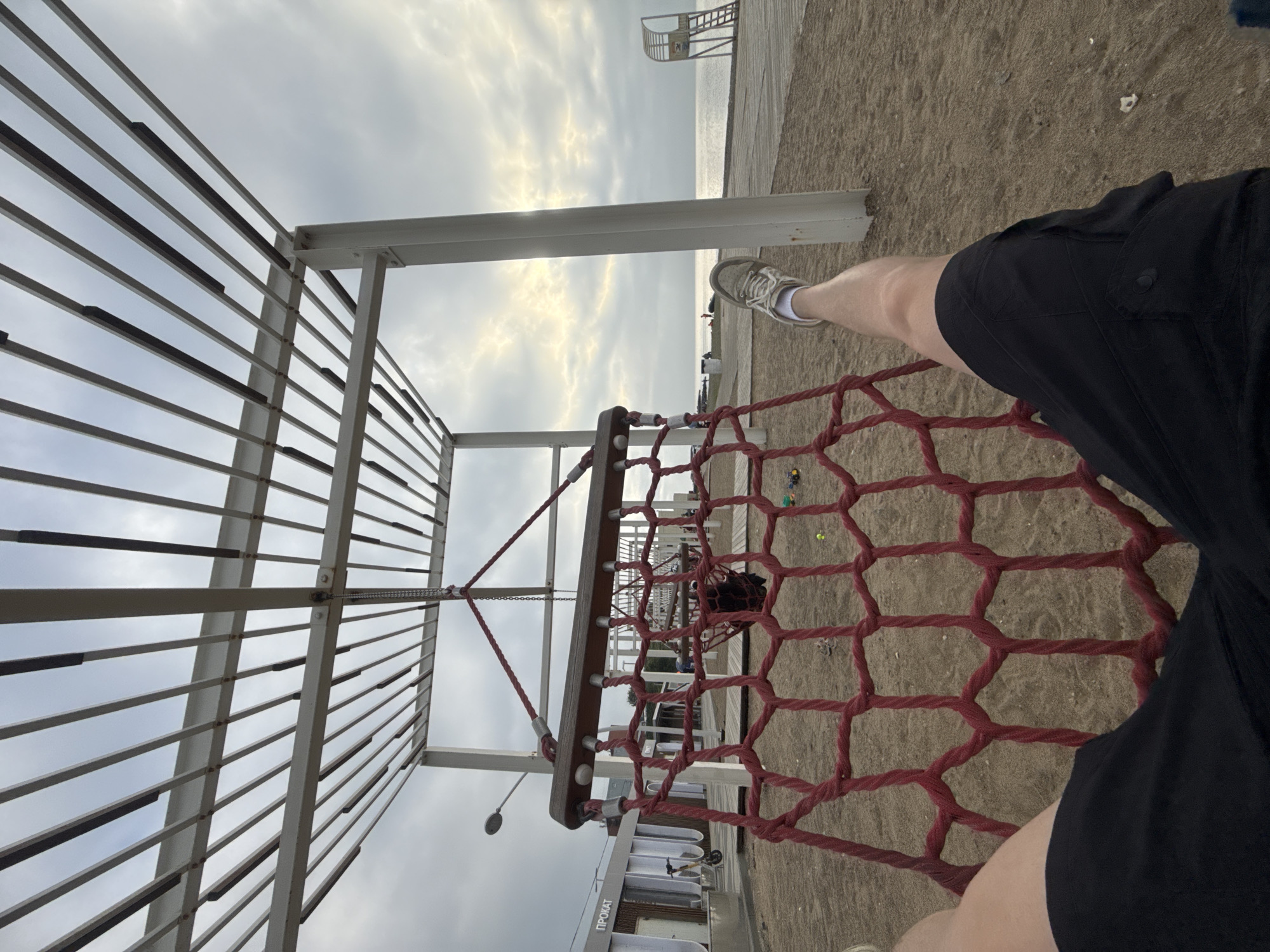 The image depicts a person's legs, clad in black shorts and a white sock with a gray sneaker, as they sit in a red rope hammock swing. The hammock is suspended from a white metal pergola structure, which is situated on a sandy beach. In the background, the ocean stretches towards a cloudy sky with hints of sunlight breaking through. Further down the beach, a wooden lifeguard tower with a ladder can be observed. To the left, a building with "ПРОКАТ" (rental) signage and a yellow electric scooter parked outside is visible. The overall environment suggests a seaside recreational area.