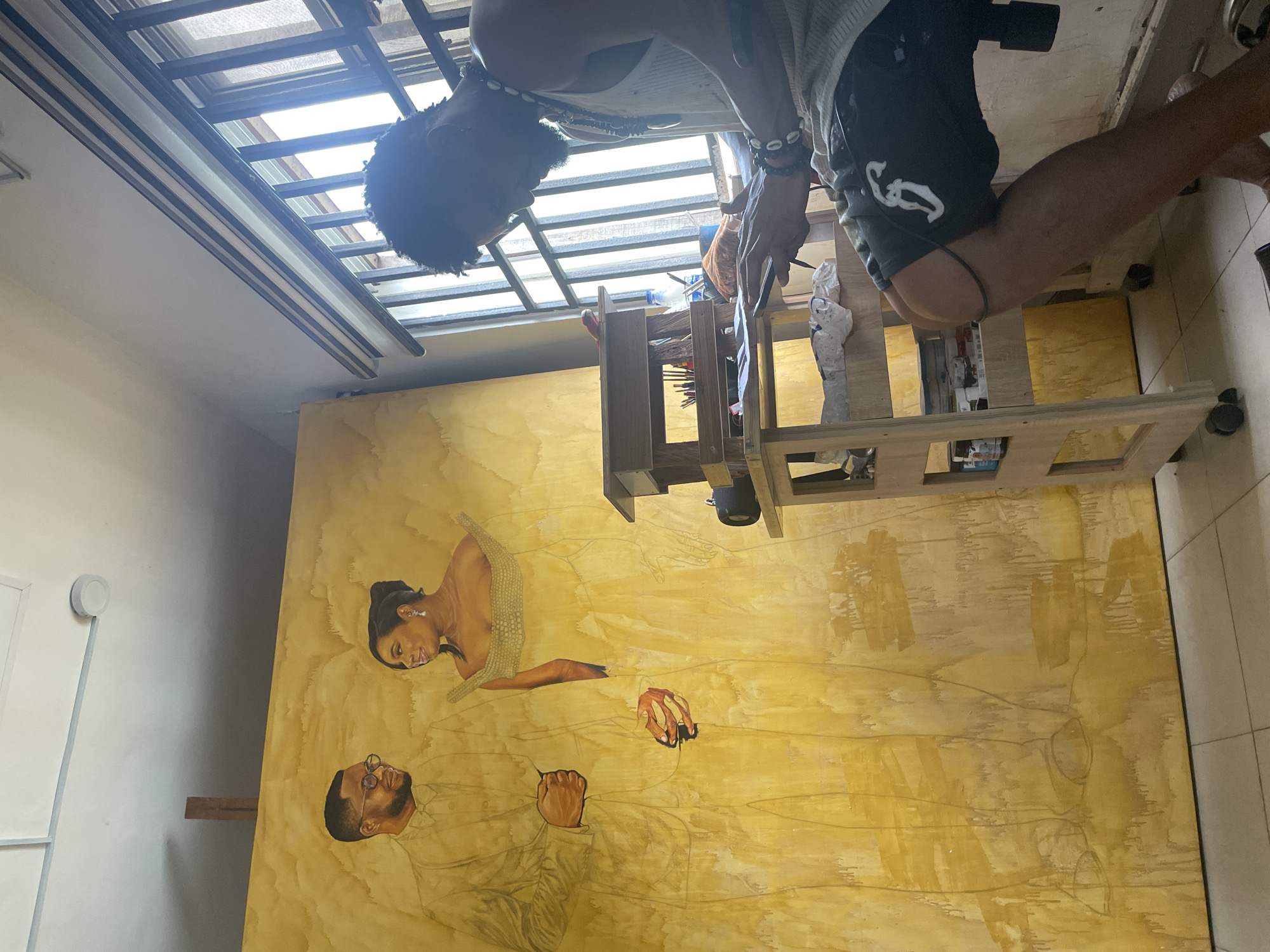 An artist is captured in a studio in Ado, Nigeria, meticulously working on a large, unfinished portrait of a couple. The artwork, rendered in warm, golden hues with visible brushstrokes and drips, depicts a man in a suit and a woman in an ornate dress. The artist, seated on a stool and partially obscured by his work, is focused on his phone, which he holds in his hands. He is dressed in a tank top and shorts, adorned with necklaces and bracelets. To his right, a multi-tiered wooden cart overflows with art supplies, including brushes, paints, and bottles. A window with metal bars and blinds provides natural light to the space. The studio floor is tiled, and the walls are a light color, contrasting with the vibrant canvas.