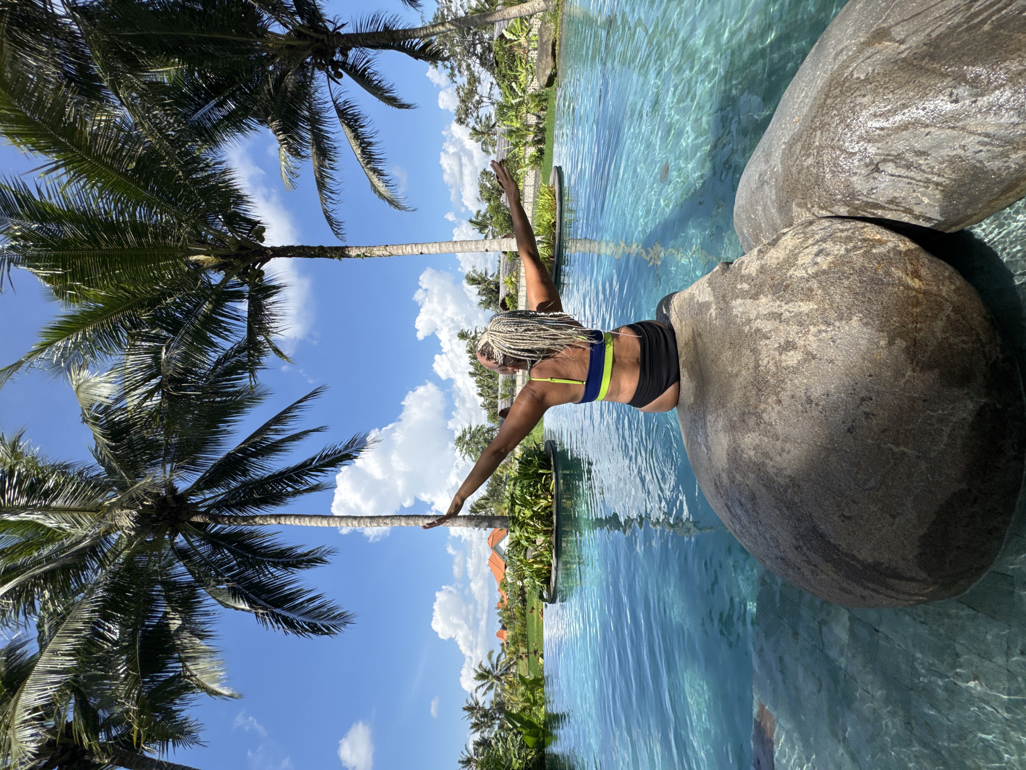 A woman stands on rocks next to an outdoor pool in Peliatan, Indonesia. The woman is tan with long blond hair in braids and is wearing a black and lime green swimsuit. She has both arms outstretched. Two palm trees are visible on the left, reaching into a blue sky with fluffy white clouds. Other trees and greenery are visible in the background near some buildings. The pool water is clear and blue.
