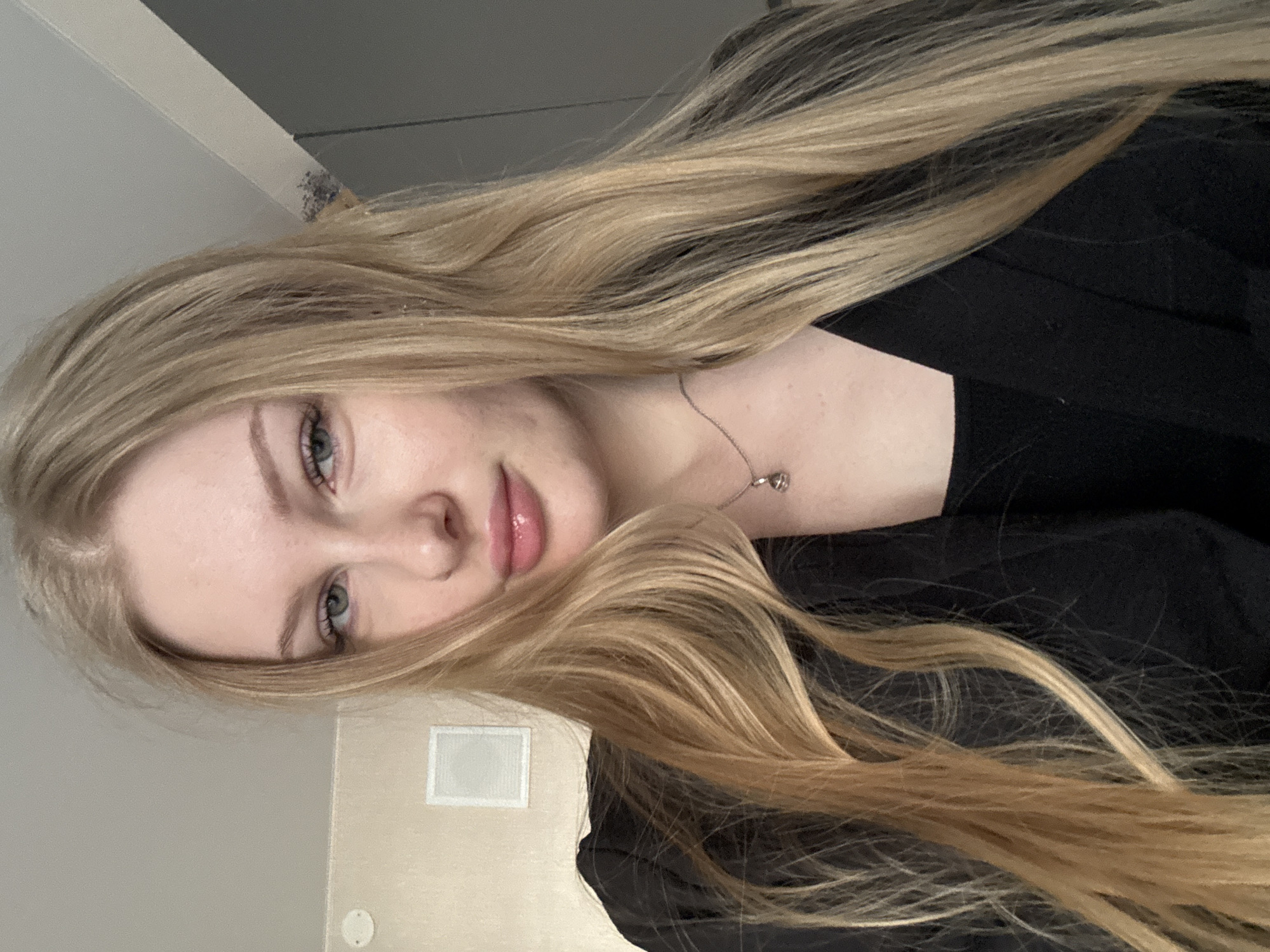 A close-up, horizontally oriented image shows a fair-skinned woman with long, blonde hair. She is wearing a dark-colored garment. A silver necklace adorns her neck. The background reveals a neutral-colored wall, an electrical outlet, and a rectangular vent. There is no indication of a location in the city of Perm, Russia.