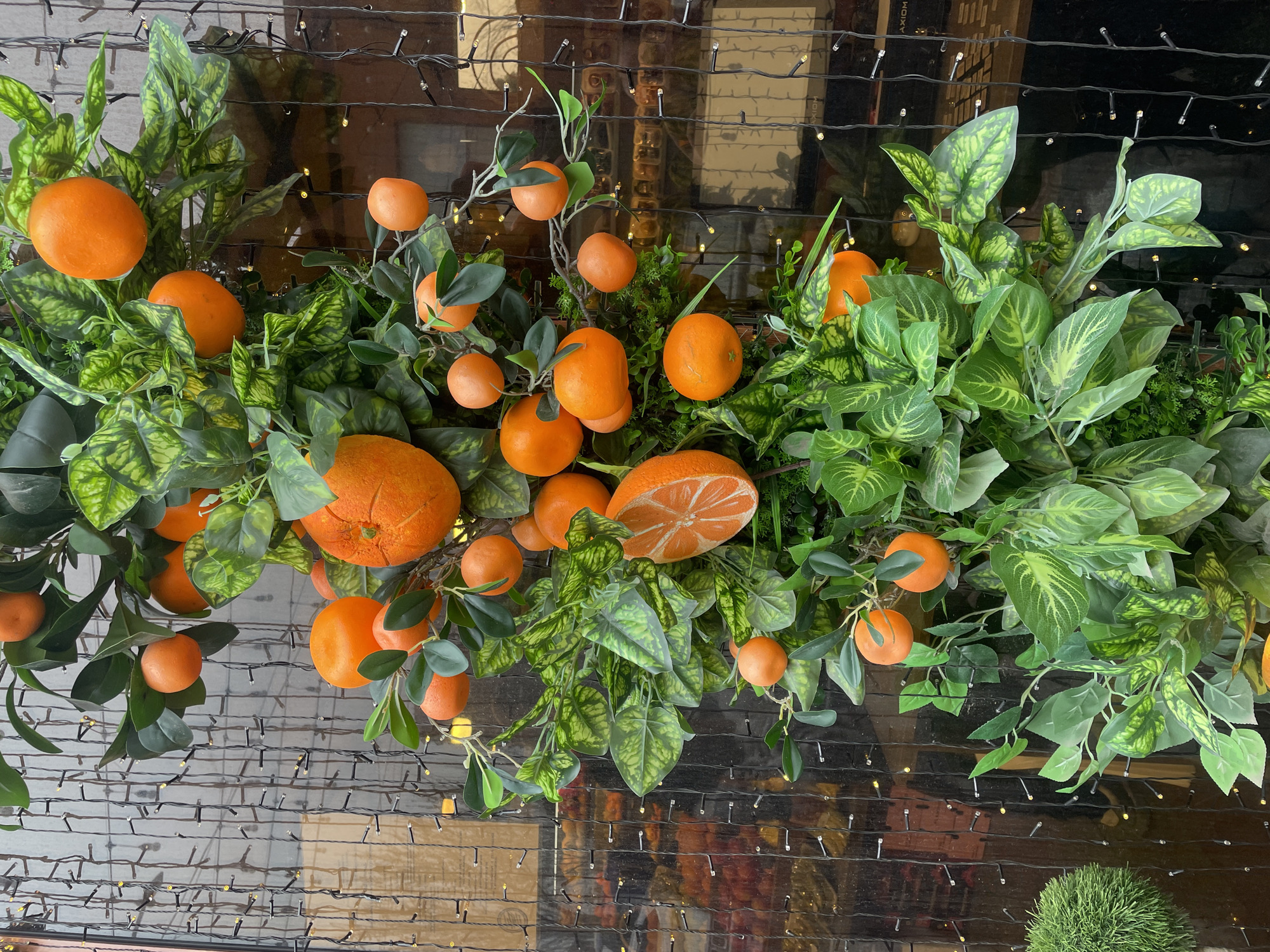 The image presents a vertical decorative display composed of artificial orange fruits and green foliage, positioned against a reflective glass surface in Yerevan, Armenia. The display features numerous artificial oranges, ranging in size, with most appearing whole and spherical. Two oranges are depicted as cross-sections, revealing an artificial segmented interior. The fruits are interspersed among artificial green leaves of varying shades and textures, simulating a natural branch structure. Behind this arrangement, vertical strands of warm-white LED string lights are strung across a dark, reflective glass panel. The glass reflects a blurred interior environment, showing indistinct shapes, colors, and light sources, indicative of a commercial space. A small, spherical topiary-like element is partially visible on the bottom left. No individuals are present in the scene.