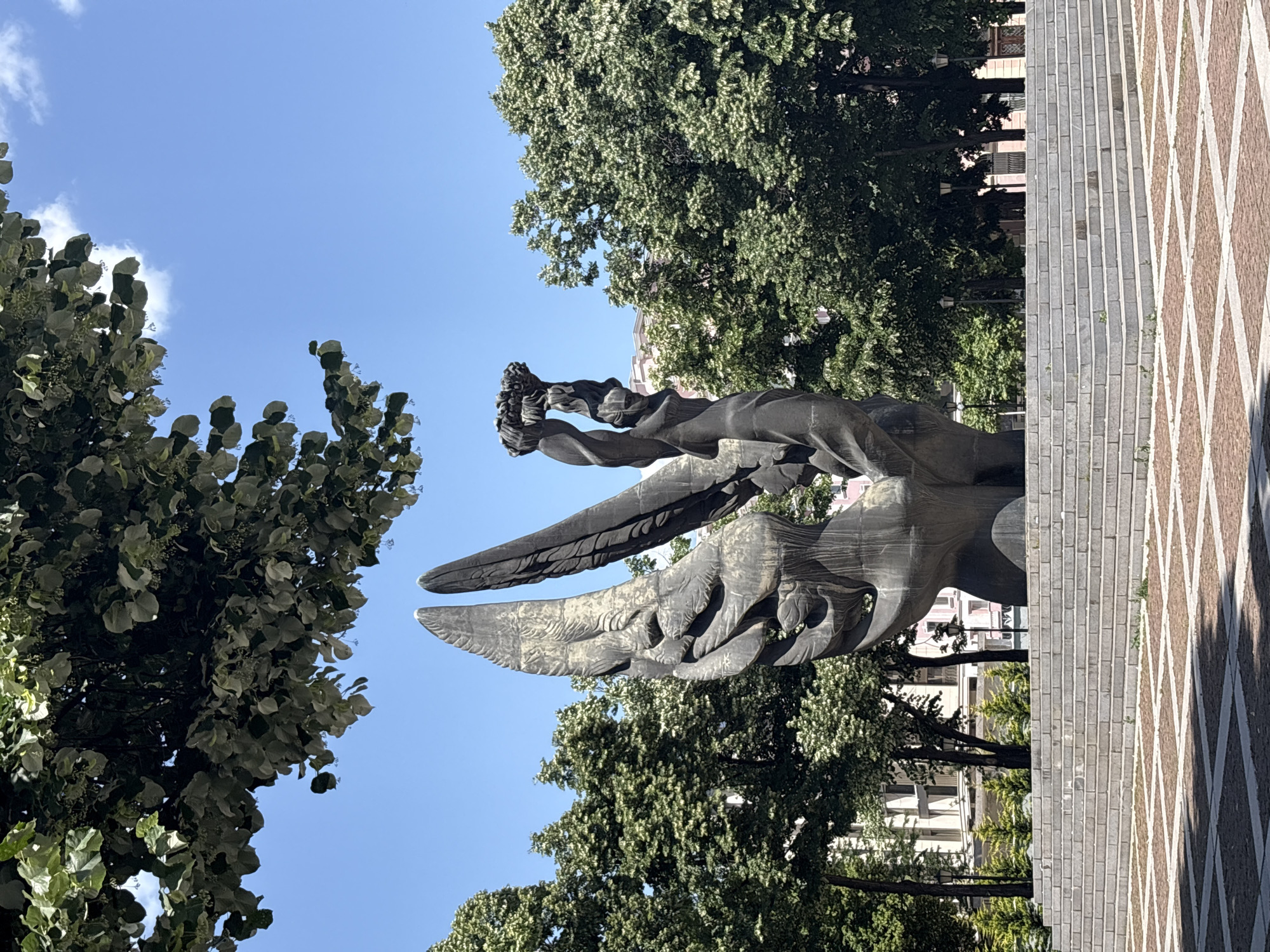 The image depicts the Alyosha Monument in Plovdiv, Bulgaria, from a low-angle perspective. The monument consists of a winged figure, seemingly made of dark metal or stone, reaching upwards with its arms. The statue is positioned against a backdrop of trees and a clear blue sky. To the right of the monument, a paved area with a geometric pattern is visible. Some buildings can be seen in the background, between the trees.