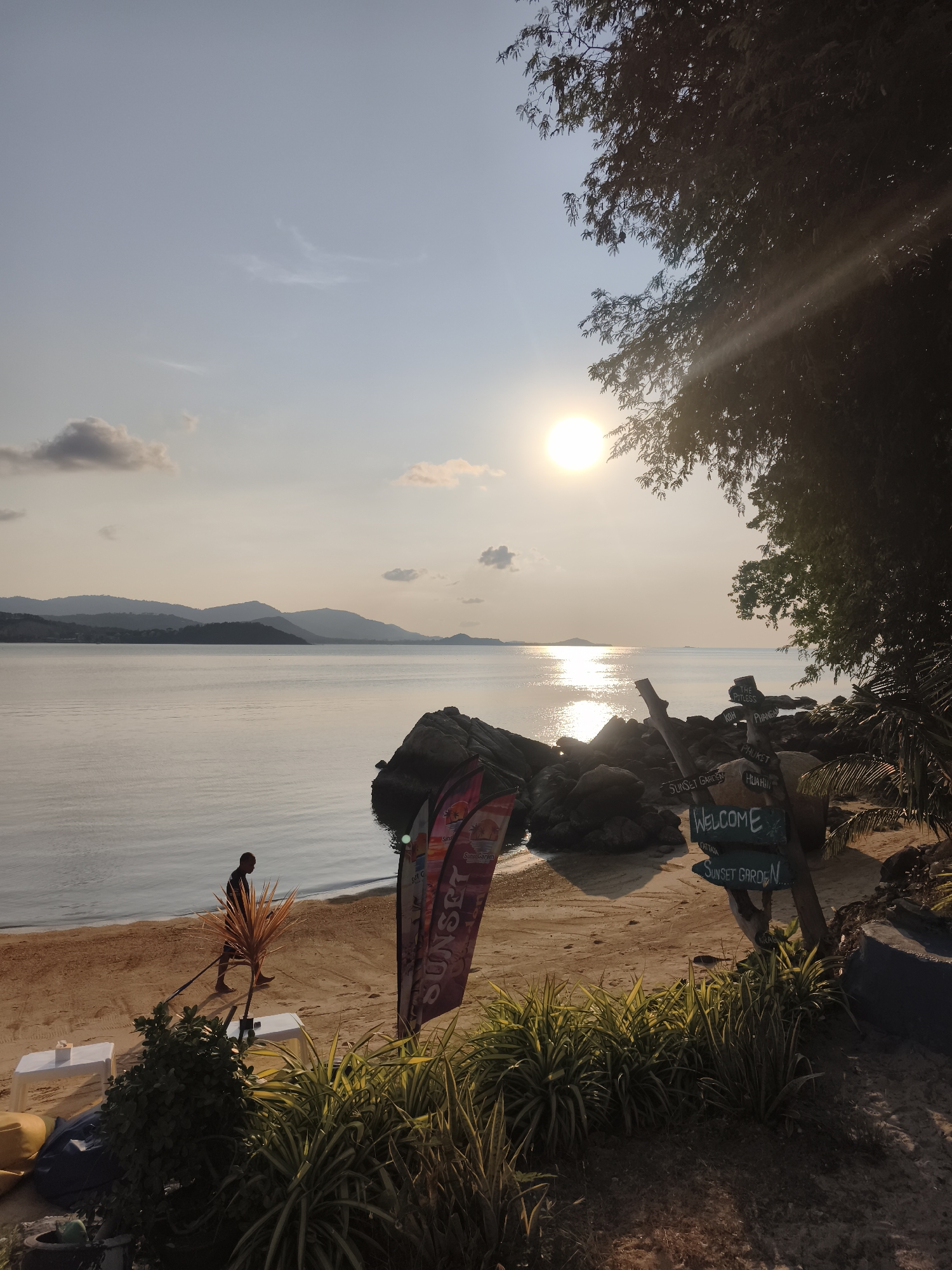 A serene coastal scene unfolds, possibly at Sunset Garden in Baan Plai Laem, Thailand, during late afternoon. The sun, a bright orb, casts a golden sheen across the calm water. In the distance, silhouetted islands or mountains are visible. A man walks along the sandy beach with what appears to be a rake and a potted plant with long thin leaves, leaving footprints in the sand. To the right, the view is partially obscured by a dark tree and a collection of rocks. A wooden signpost bearing the word "WELCOME" and several place names, including "KOH PHANGAN," is nestled amongst the rocks. The scene also includes green shrubbery and a "Sunset" banner. A couple of white tables are set near the ground. The atmosphere is one of peace and tranquility.