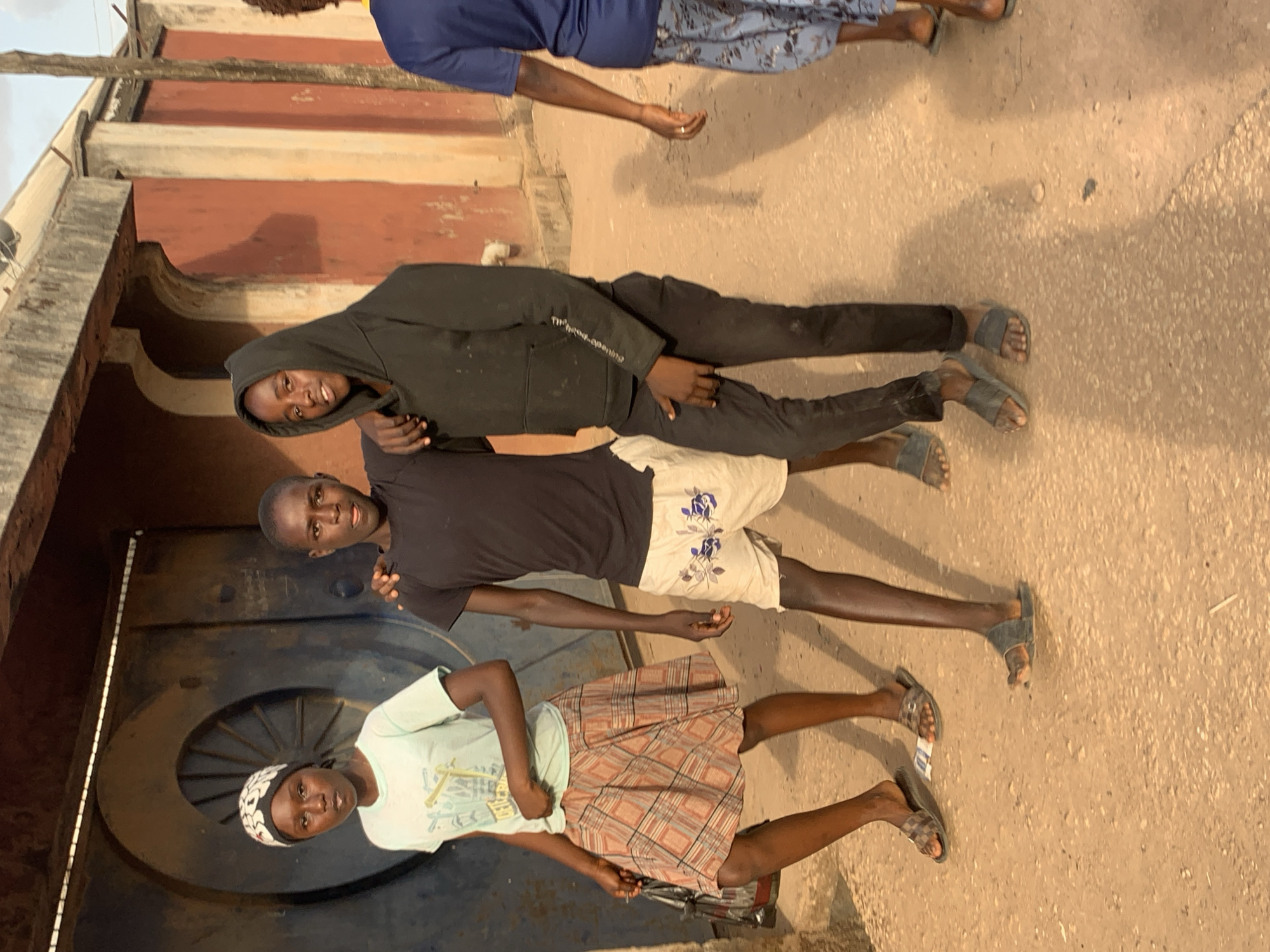 Three individuals stand outdoors on a textured, concrete-like surface. The setting appears to be near a building, evident from the brick facade and a large dark door visible in the background. The first individual, positioned closest to the camera, wears a light green t-shirt, a patterned skirt, and a head covering. The middle individual is in a black t-shirt and patterned shorts. The third individual, standing behind the middle one, is wearing a black hoodie and pants. The photo is taken in the city of Jos, Nigeria.