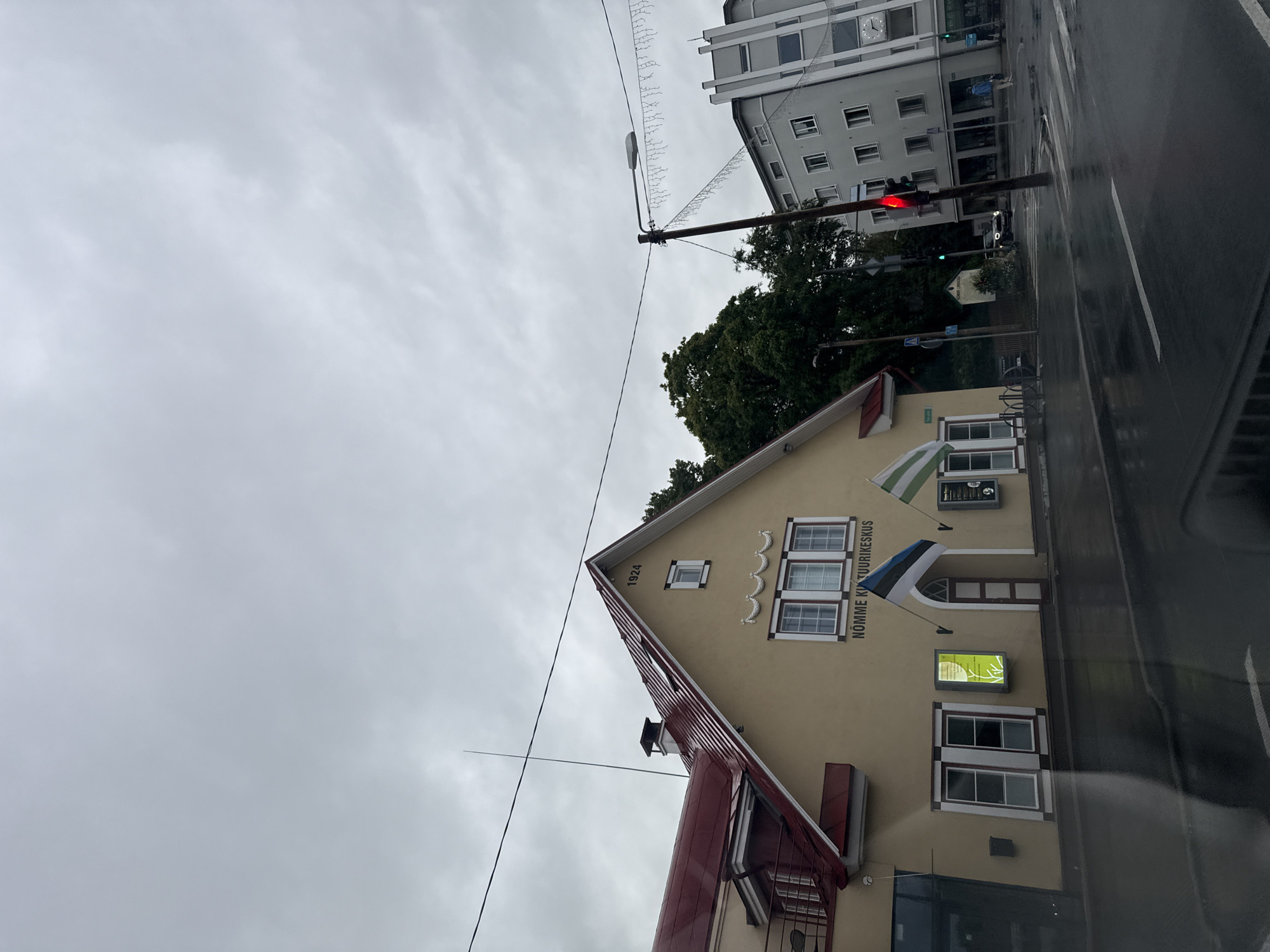 The image shows a street scene in Tallinn, Estonia, with a cloudy sky overhead. In the foreground, a wet asphalt road reflects the muted light. A traffic light is visible, showing a red signal. To the left of the road is a tan-colored building with a red roof. Above its entrance, the text "NÕMME KULTUURIKESKUS" is visible. An Estonian flag is displayed prominently near the entrance. Further to the right, a modern, grey multi-story building stands. Trees are present in the background. The scene appears to be an urban environment on a potentially rainy day.