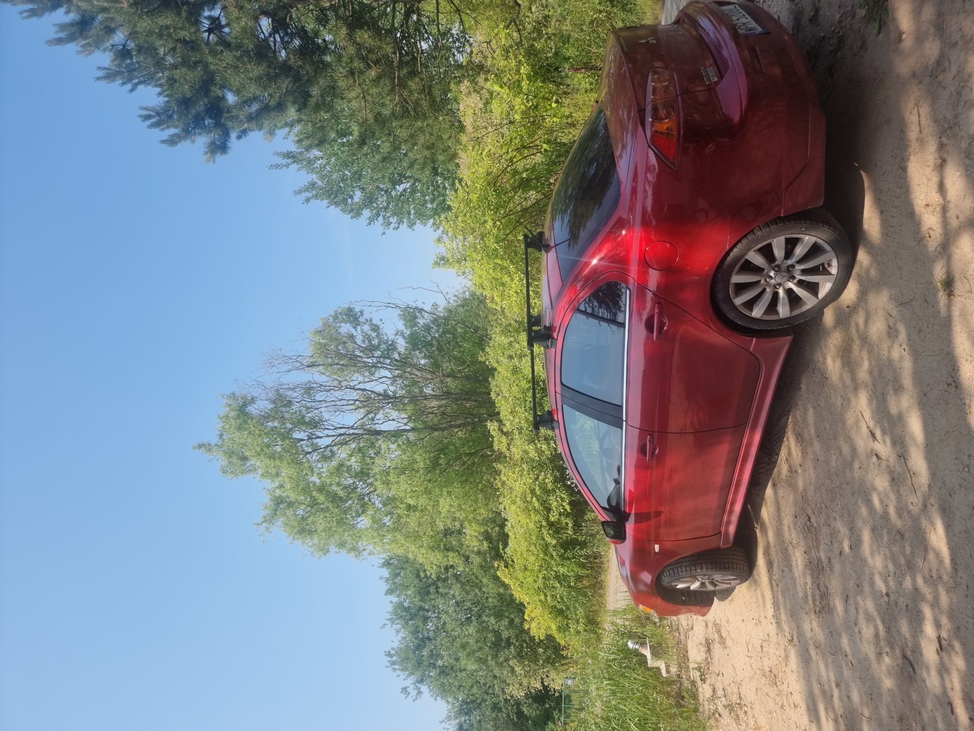 A red Mitsubishi Lancer sedan is parked on a dirt road in a rural area, possibly in Szczaki, Poland, on a sunny day. The car has a roof rack and is angled towards the left side of the frame. Lush green trees and bushes surround the road, with a clear blue sky overhead. The ground is sandy and dry, with shadows cast by the trees. There are no people visible in the image.
