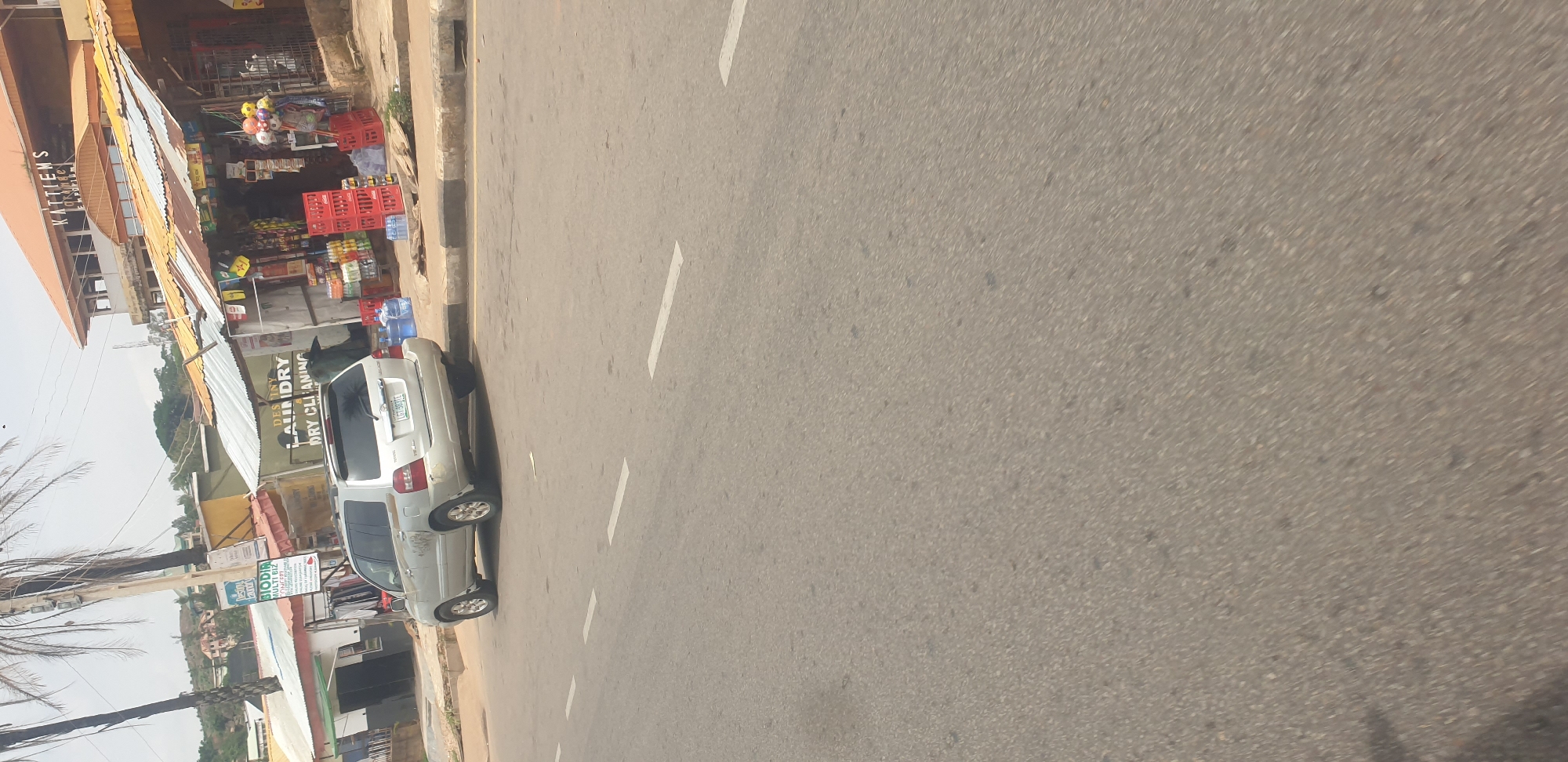 The image shows a street scene in Jos, Nigeria. In the foreground, there is a paved road with white lane markings. On the right side of the road, there is a silver Toyota Highlander SUV parked, with its rear facing the viewer. The vehicle has a license plate that reads "LGT 983TE". Beside the car and behind it, there are several shops with corrugated iron roofs. One shop displays "DESTINY LAUNDRY DRY CLEANING" on its facade. Another shop, labeled "KATTIEMS PARADISE EMPORIUM", is visible further down. Red plastic crates filled with bottles and other items are stacked outside the shops. The environment appears to be a commercial area in an urban setting, with buildings and palm trees visible in the background. The sky is overcast.