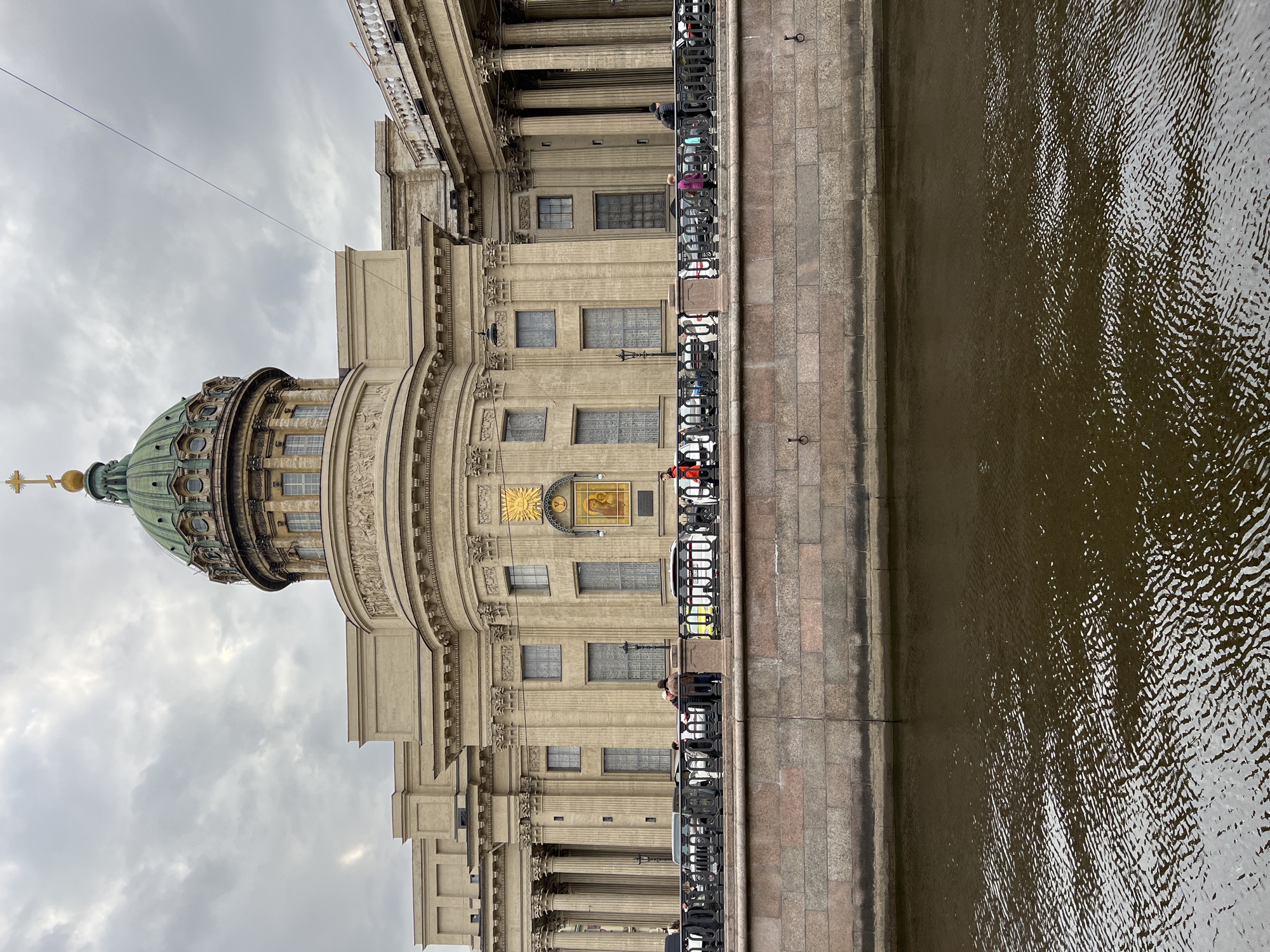 The image displays a grand neoclassical building, identified as Kazan Cathedral in Saint Petersburg, Russia, under an overcast sky. The photograph is taken from across a body of water, likely the Griboyedov Canal, looking towards the building.
In the foreground, occupying the bottom right portion of the frame, is a dark, rippled body of water, reflecting the muted sky with patterns of small waves. Adjacent to the water, a stone embankment made of grey and reddish-brown blocks runs horizontally across the lower part of the scene. It is topped with an ornate black metal railing, behind which several people are visible. Some individuals are standing by the railing, others appear to be strolling along the embankment. One person wears a distinctive red jacket.
The Kazan Cathedral dominates the left and central parts of the image. Its imposing facade is constructed from light beige stone, characterized by numerous tall classical columns with intricate capitals and detailed friezes and cornices. A prominent feature is the cathedral's large green dome, which rises towards the upper left, crowned with a golden cross. On the central facade, above a row of windows, a golden sunburst motif is depicted. Below this, a rectangular golden frame encloses a religious icon, though its specific details are not clearly discernible. Multiple windows with grid patterns are visible across the building's facade.
The sky is uniformly overcast with soft grey clouds, indicating a cloudy day with diffused light. There is no direct sunlight. The scene conveys a calm, somewhat subdued atmosphere, with the historic architecture serving as the primary subject against the tranquil backdrop of the water and sky. No specific textual information beyond the general imagery of the icon is clearly readable.