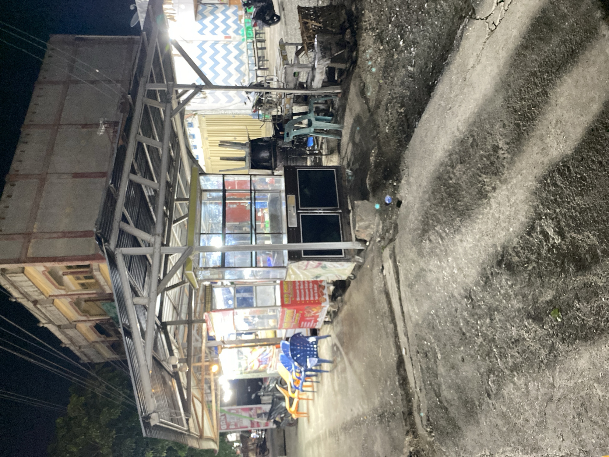 The image shows an outdoor street scene at night in Banda Aceh, Indonesia. The perspective is from ground level, looking upwards towards a row of roadside stalls or shops. The scene is illuminated by artificial lighting, indicating it is nighttime.
The main subjects are the structures of the stalls, which appear to be made of metal frames and corrugated metal sheeting, with some having translucent coverings. There are tables and chairs set up outside some of the stalls, suggesting a dining or gathering area. Some chairs are plastic, in colors like blue, yellow, and orange. Several stalls have illuminated signage, with some displaying food items or advertisements. A motorcycle is parked in the background near one of the stalls. The ground appears to be asphalt or concrete, with some debris and unevenness. The overall setting is a busy, informal commercial area.
Visible text on the signs includes what appears to be "GAN" and a red sign with white text, possibly a menu or advertisement. The lighting suggests it is not raining. The overall atmosphere is one of a lively night market or street food area.