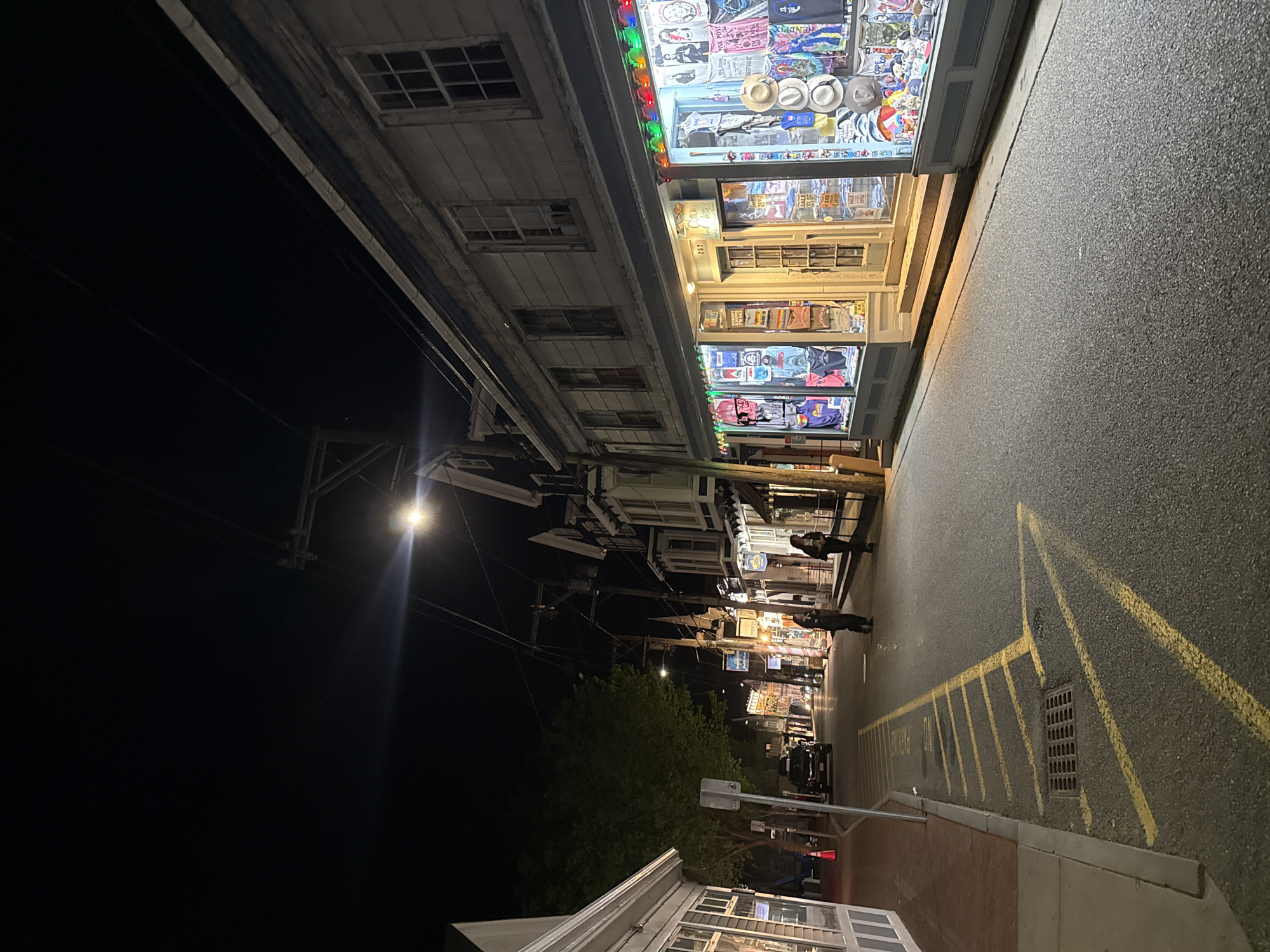 The image depicts a nighttime street scene in Provincetown, Massachusetts, United States. The view is along a street lined with buildings, their facades featuring display windows illuminated with merchandise. Two pedestrians are visible, walking along the sidewalk. The street itself has yellow parking lines painted on its surface. A street lamp is positioned to the left, and a vehicle is parked further down the street. The sky is dark, and trees are present, adding to the scene's urban and natural elements.