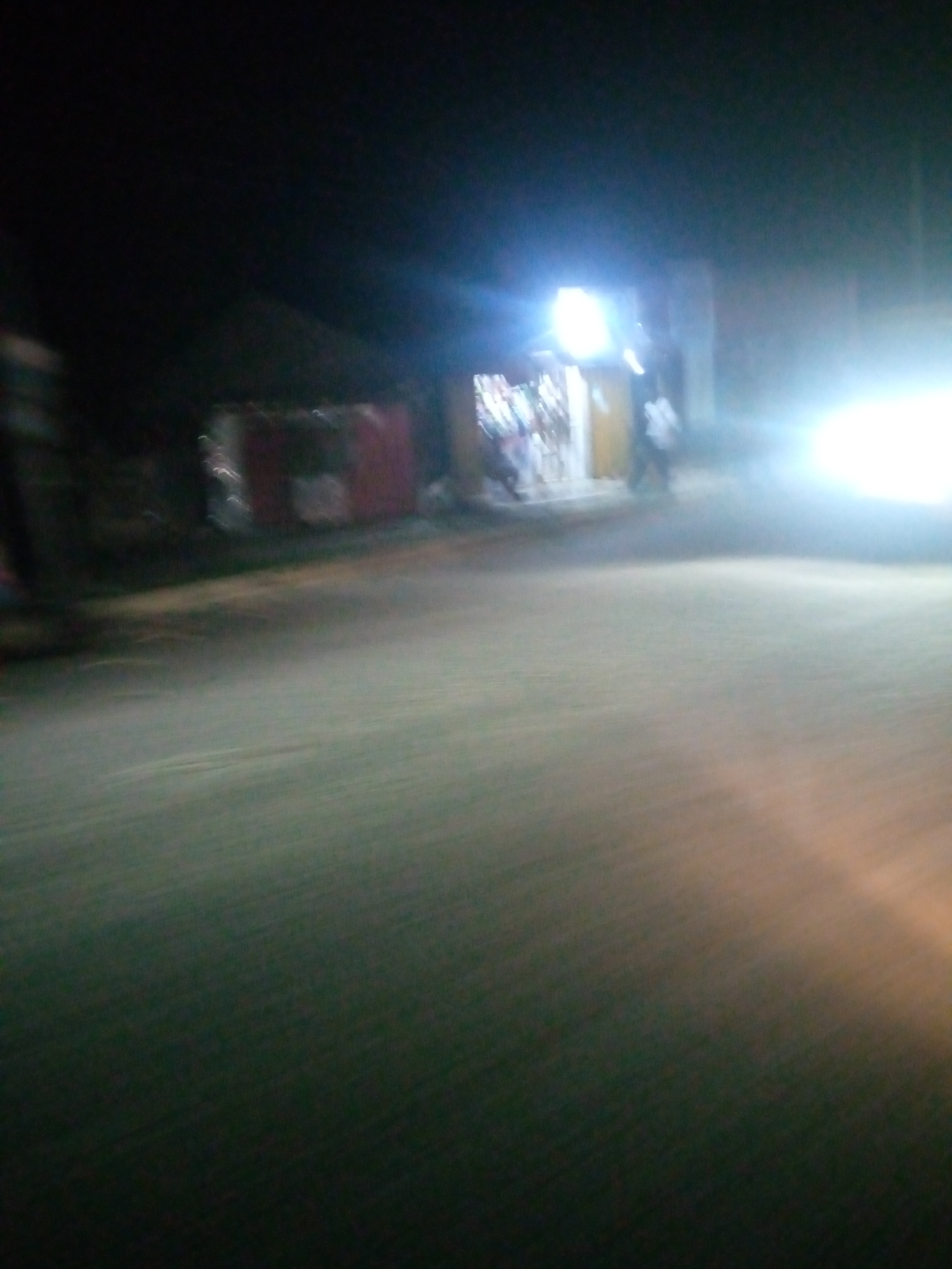 The image is a blurry, low-light photograph taken at night, likely from a moving vehicle. It depicts a street scene in Jalingo, Nigeria, with several small shops or market stalls lining the side of the road. Bright lights, possibly from a vehicle's headlights or streetlights, illuminate parts of the scene, creating significant glare and reducing overall visibility. The ground appears to be a paved road or dirt track. There are indistinct figures of people in the background, one of whom appears to be standing near a brightly lit stall. The overall impression is one of a busy, albeit dimly lit, urban environment at night.