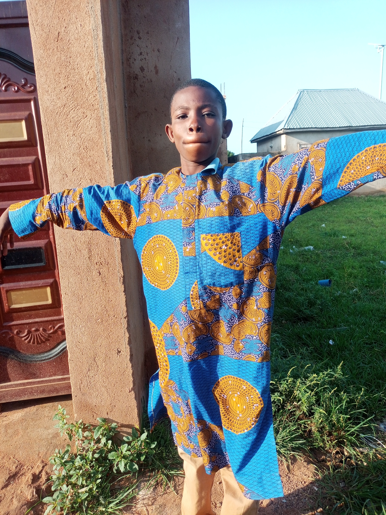 A young boy stands in front of a tan-colored wall, his arms outstretched. He wears a blue and gold patterned tunic and light brown pants. A red and gold patterned door is visible to his left. The background consists of green grass, a building with a silver roof, and a clear blue sky. The scene appears to be outdoors in a residential area.