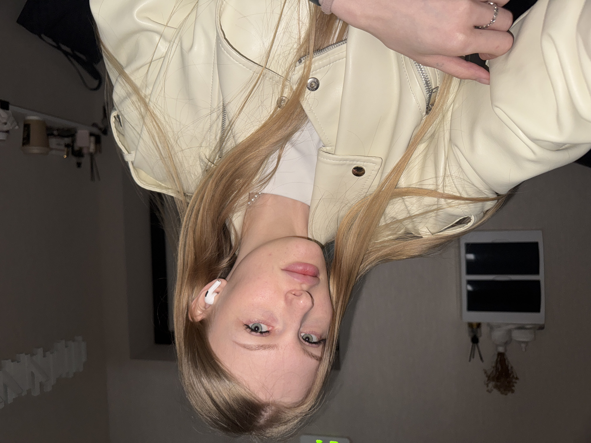 The image features a young woman with long, straight, light blonde or light brown hair, looking directly at the camera. She has light blue eyes and a neutral to slightly serious expression. She is wearing a light cream or off-white jacket, which appears to be made of faux leather, featuring a front zipper and subtle quilted detailing on the shoulders. A white wireless earbud is visible in her left ear, and a silver ring with a patterned band is worn on a finger of her right hand.
The setting is an indoor space, likely a room, characterized by soft, possibly artificial lighting, suggesting evening or night. The background is blurred, but several objects can be identified. To the viewer's right, a white rectangular unit with two dark panels or screens is mounted on a wall, with decorative tassels or keychains hanging beneath it. To the viewer's left, a dark, indistinct object (possibly an electronic screen) and a shelf or rail holding various small items, such as jars or bottles, are visible. The activity captured is a posed portrait. No discernible text is visible in the image. This photo was taken in Perm, Russia.