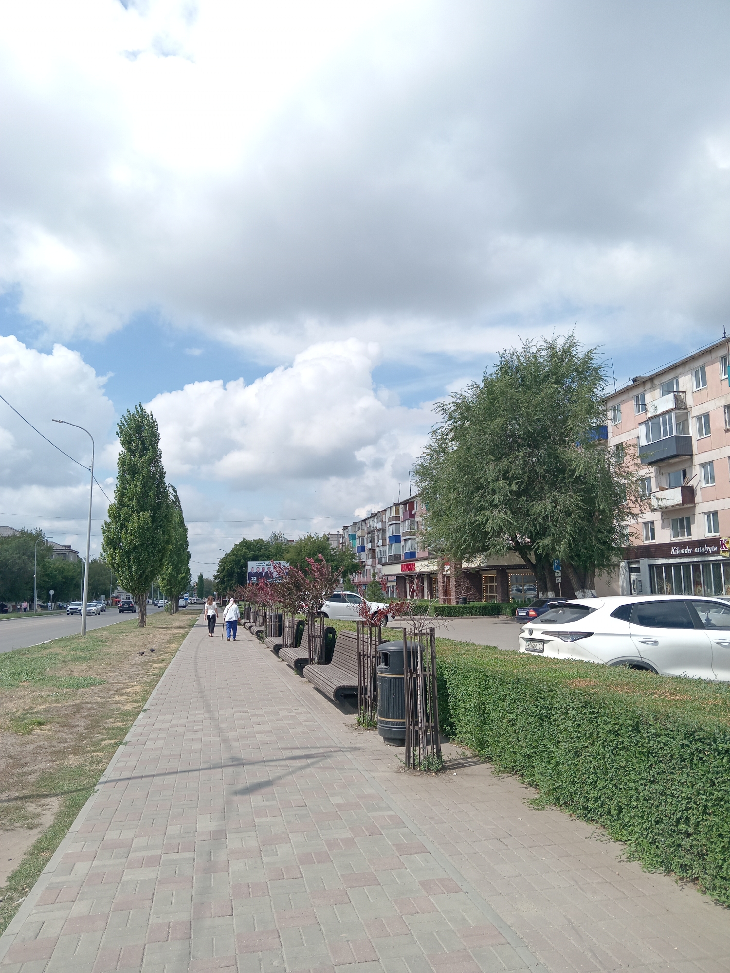 The image depicts a street scene in Rudny, Kazakhstan. The perspective is from a sidewalk paved with rectangular bricks, looking down the street. On the left, a road with several cars driving is visible, separated from the sidewalk by a strip of dry grass and then a well-maintained green lawn. Tall, slender trees line the edge of the sidewalk.
The sidewalk itself is wide and features several wooden benches and dark cylindrical trash receptacles. Further down the sidewalk, two women are walking away from the camera. One is wearing a white headscarf and a light-colored outfit, leaning on a cane, and the other is in a white top and dark pants.
On the right side of the street, there are multi-story apartment buildings with balconies. Some of these buildings have shops or businesses on the ground floor, indicated by signs such as "Boutique" and "Kilemdar ortalyqta." A white car is parked in front of one of these buildings. A neatly trimmed green hedge runs along the edge of the sidewalk on the right, separating it from the parked cars and buildings. The sky above is partly cloudy with white cumulus clouds scattered across a blue background. The overall impression is that of a typical urban street in fair weather.
