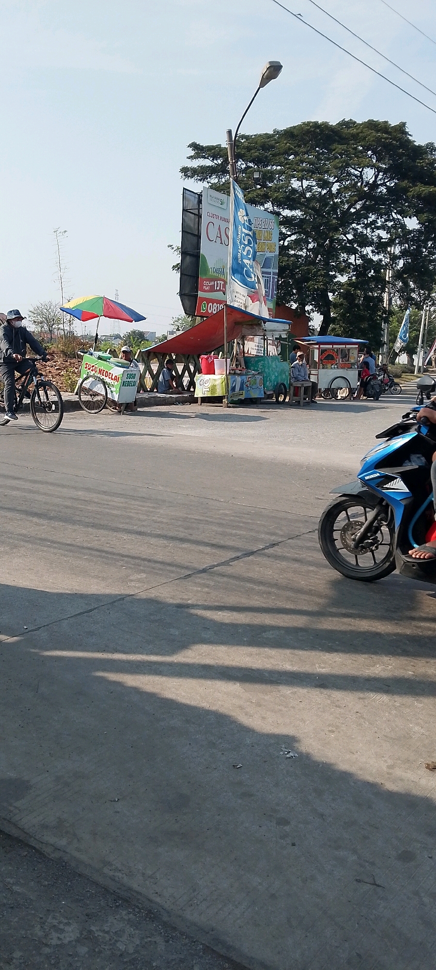 The image captures a street scene in Sriamur, Indonesia, on a sunny day. The foreground is dominated by a concrete road with distinct tire tracks and the dappled shadows of overhead trees. On the right side of the road, a blue and black motorcycle is partially visible, with its front wheel and part of the rider's leg wearing a sandal. The middle ground features a row of street vendors operating from carts and makeshift stalls under a canopy. One vendor cart has a blue and white color scheme with text that is partially legible. To the left of the vendors, a person on a bicycle wearing a hat and mask rides on the road, pulling a cart with colorful umbrellas and signage that reads "SUSU KEDELAI" (Soy Milk) in green lettering. Several other individuals are seated or standing near the vendor stalls. In the background, a large billboard for a residential cluster named "CASSIAS" is prominently displayed, with trees and utility poles visible against a clear sky.
