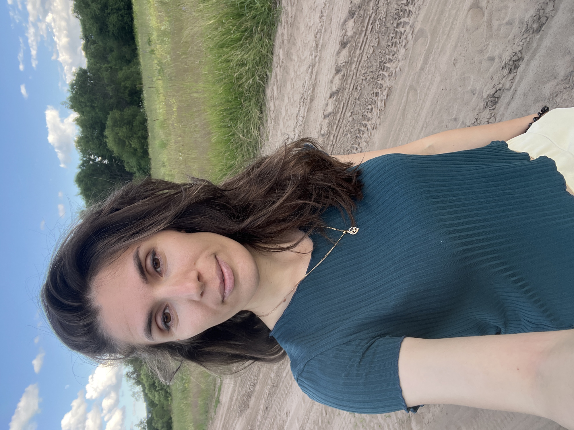 A woman with long, dark, wavy hair is taking a selfie outdoors on a dirt road. She is wearing a dark teal, short-sleeved, ribbed top and light-colored bottoms. She is also wearing a delicate gold necklace with a pendant. The background features a field of tall grass and wildflowers, with a dense line of green trees and a blue sky with scattered white clouds. The location appears to be rural, with no buildings or urban features visible. The ground in the foreground is a sandy or dirt path with tire tracks. The overall impression is of a natural, outdoor setting.