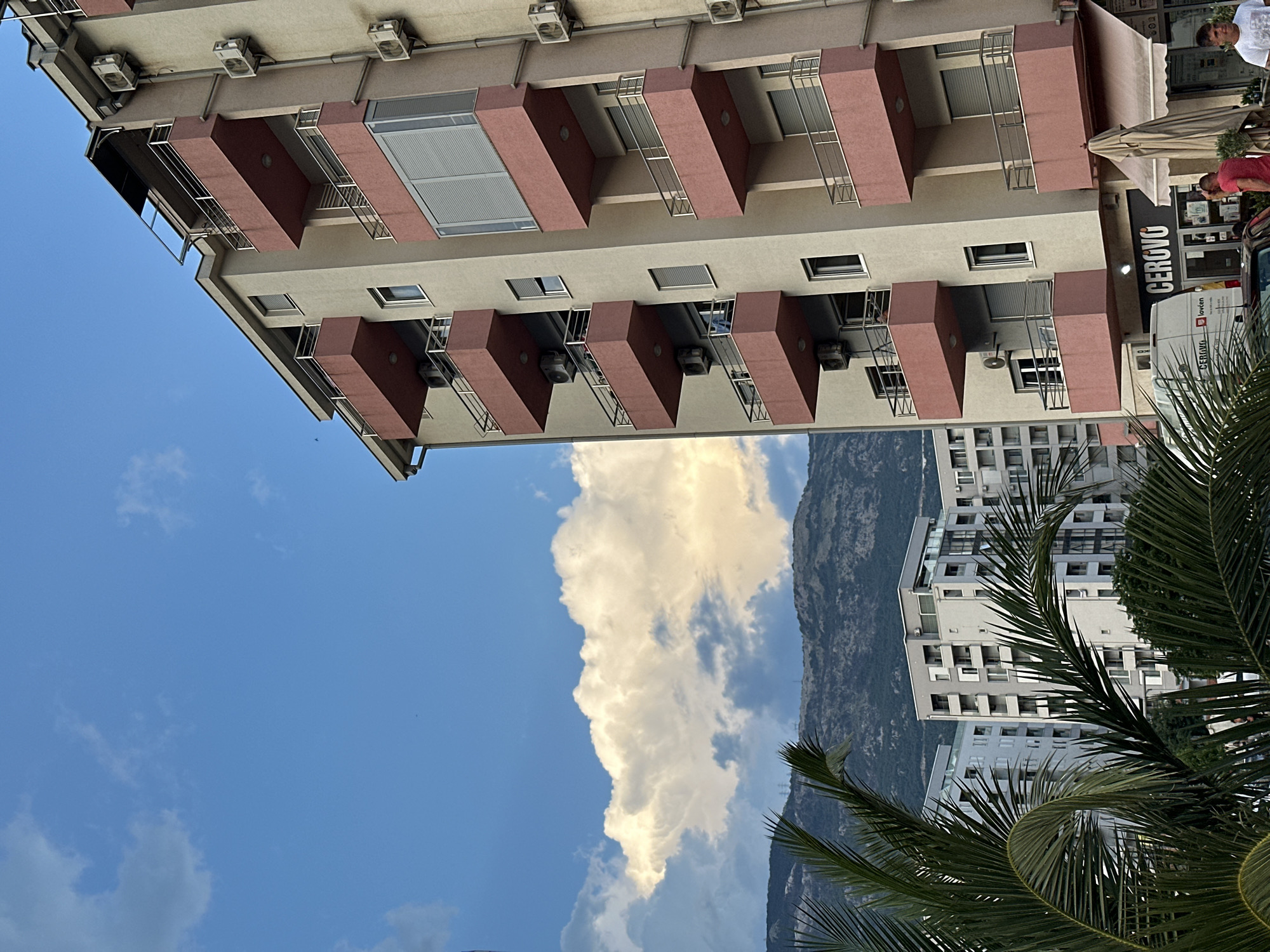 This is a vertically oriented outdoor photograph taken in Budva, Montenegro. The scene depicts buildings, clouds, and a glimpse of a mountain. In the foreground, a palm frond is visible on the right side, obscuring part of the view.
The primary subjects are two multi-story buildings. The building on the left has a light-colored facade with numerous balconies, each featuring a reddish-brown railing and a projecting rectangular structure. Several air conditioning units are visible mounted on the exterior of this building. The building on the right appears more modern, with a grid of windows and balconies.
A large, white cloud dominates the sky, catching the light and creating a dramatic contrast with the deep blue sky. A mountain range is visible in the background, its slopes appearing rugged and covered in vegetation.
There are a few people present, mostly indistinct figures. On the upper right, two men are standing near an awning. Another person is partially visible near the entrance of a shop.
A sign on the building on the right reads "CERCIO." A white van with "CERCIO" printed on its side is parked in front of the shop. The overall impression is of a sunny day, likely late afternoon given the warm light illuminating the cloud. The presence of palm trees and the mountainous backdrop are characteristic of coastal Mediterranean locations like Budva.