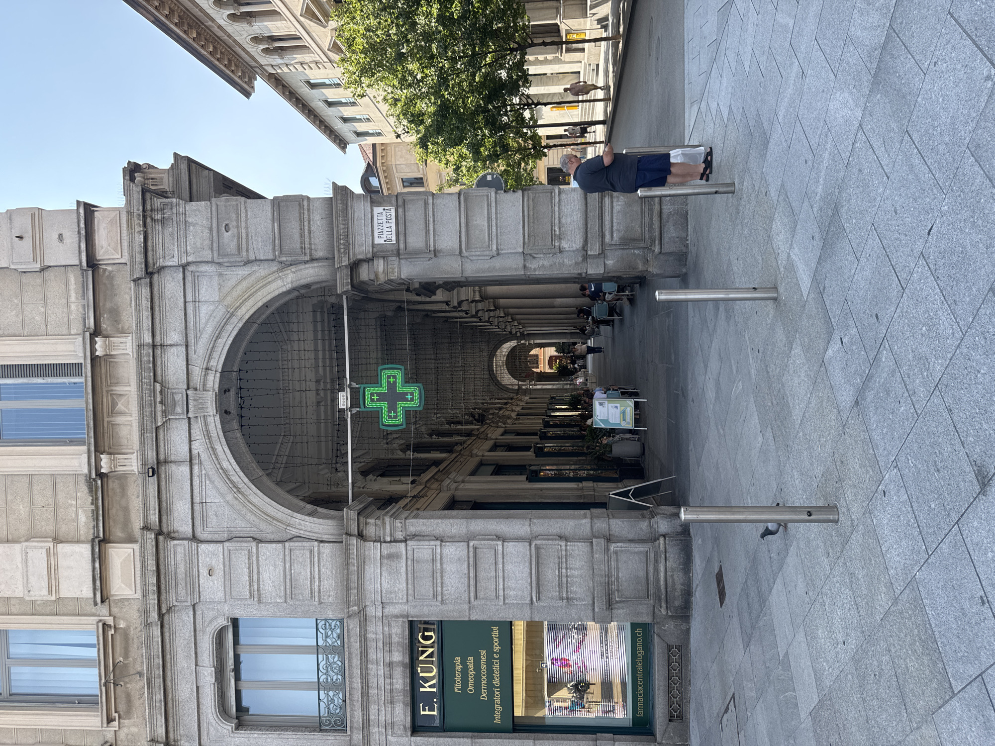 A full shot captures the exterior of a building with an arched colonnade in Lugano, Switzerland, on a sunny day. The building features a grey stone facade with classical architectural elements, including ornate window frames and layered columns. A prominent green, illuminated cross sign, typical for a pharmacy, hangs within the colonnade. To the left, a pharmacy storefront with the name "E. KÜNG" is visible, displaying services like "Fitoterapia," "Omeopatia," and "Dermocosmesi." In front of the pharmacy, a pigeon stands on the grey tiled pavement.
The colonnade extends into the distance, revealing a walkway lined with columns and archways. Several tables and chairs are set up under the colonnade, with a few patrons seated and others walking by. On the right side of the image, a street runs parallel to the building, with trees providing shade. A man wearing a dark blue t-shirt and shorts stands near a lamppost on the sidewalk, holding a white plastic bag and looking at his phone. Further down the street, other pedestrians are visible. A sign on a pillar reads "PIAZZETTA DELLA POSTA." The sky is clear and blue.